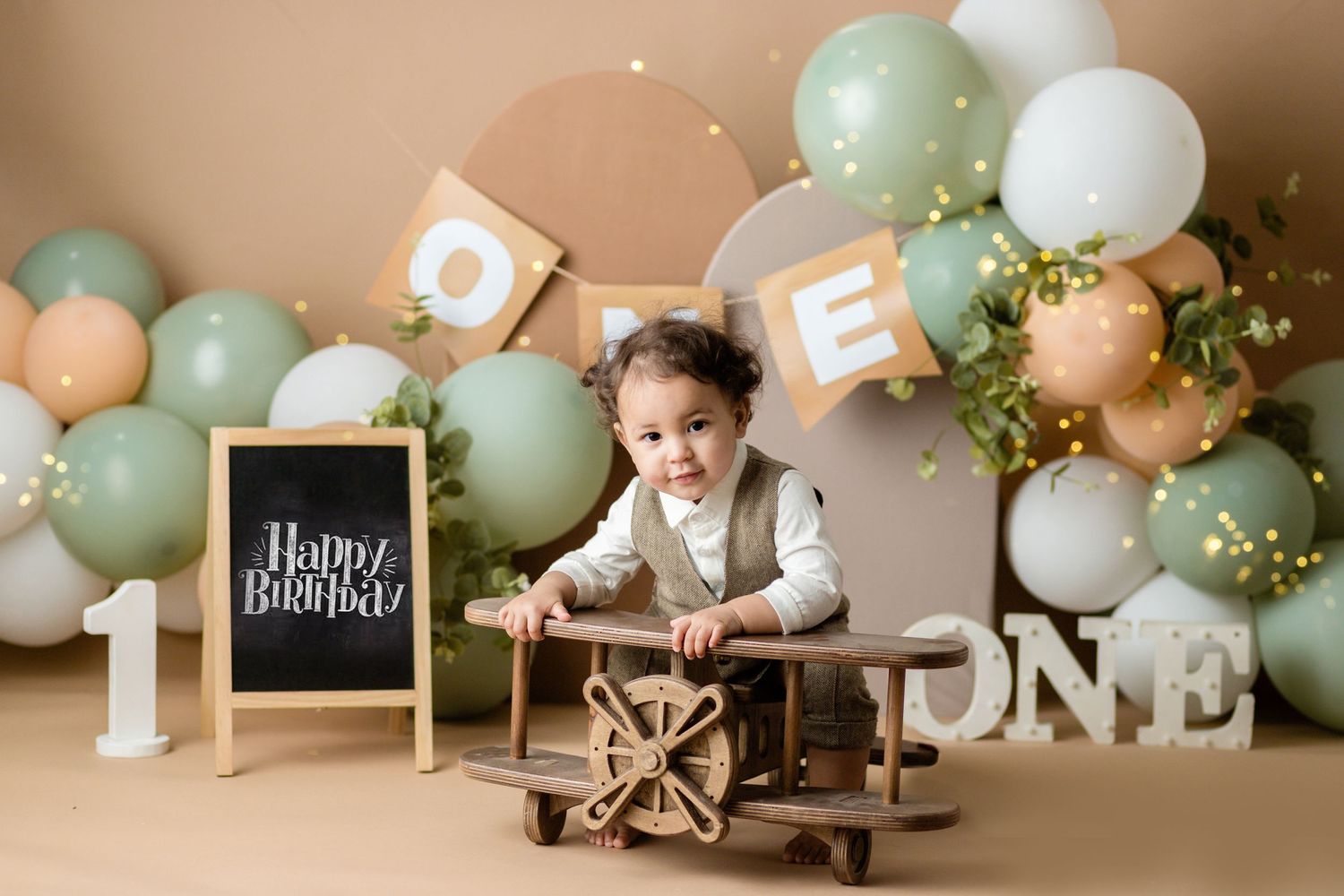 First birthday party setup with vintage wooden airplane prop against pastel balloon arch and chalkboard sign.
