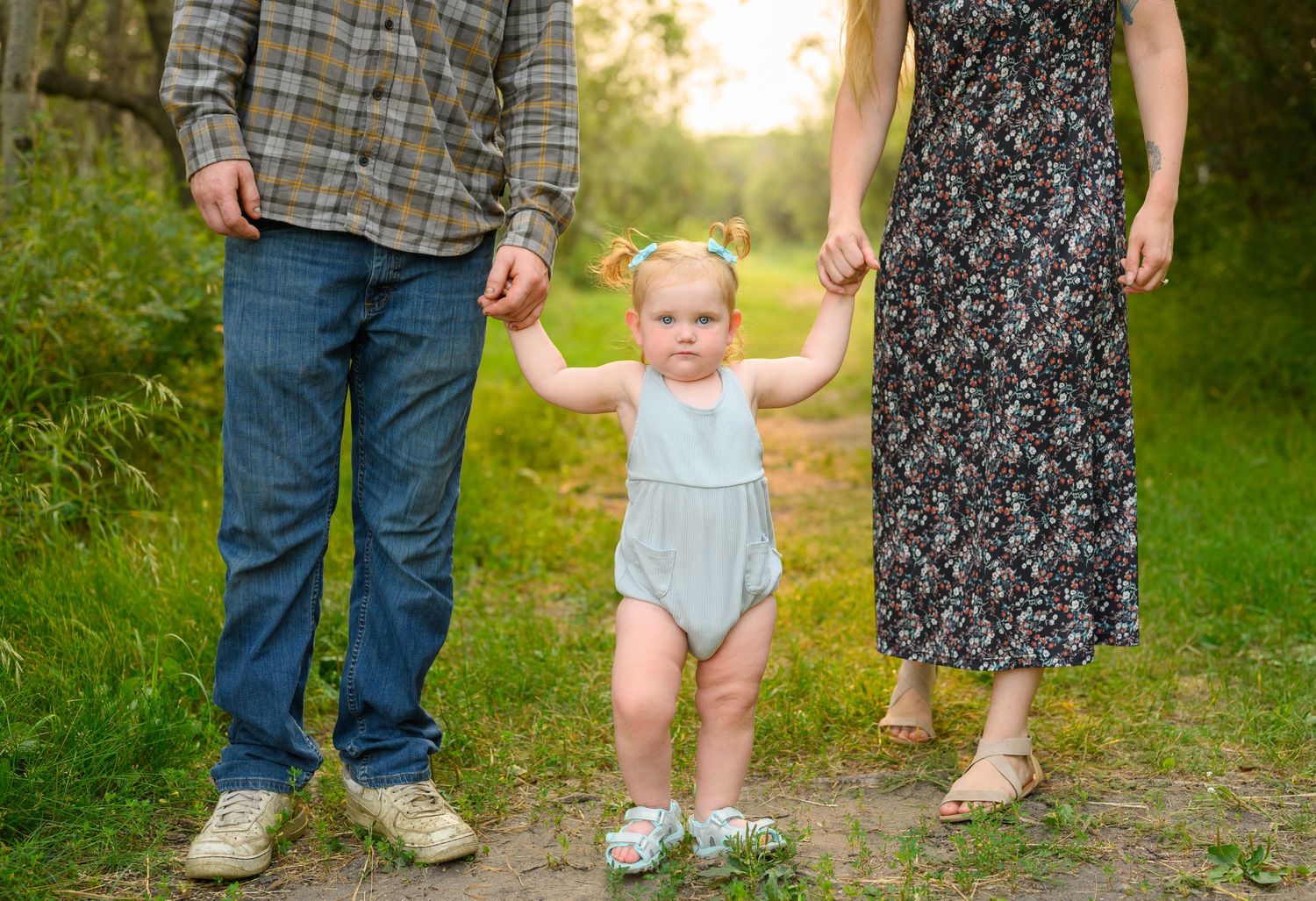 A young toddler walks hand-in-hand with parents on a forest path.