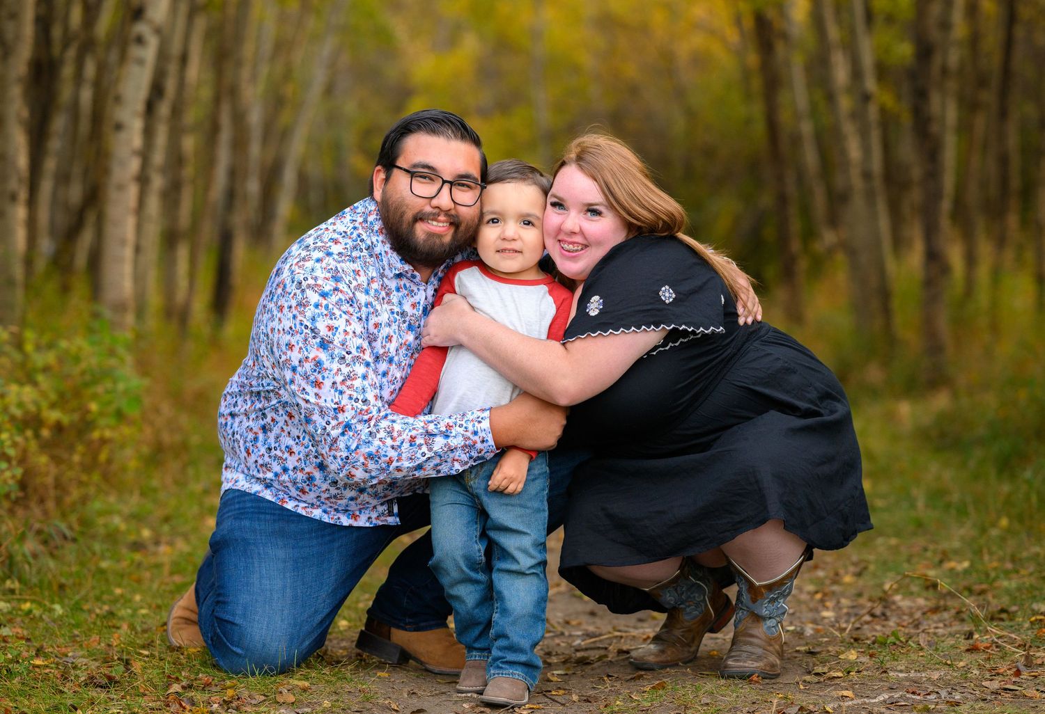 A family poses together on a tree-lined path during autumn, embracing while kneeling in fallen leaves.