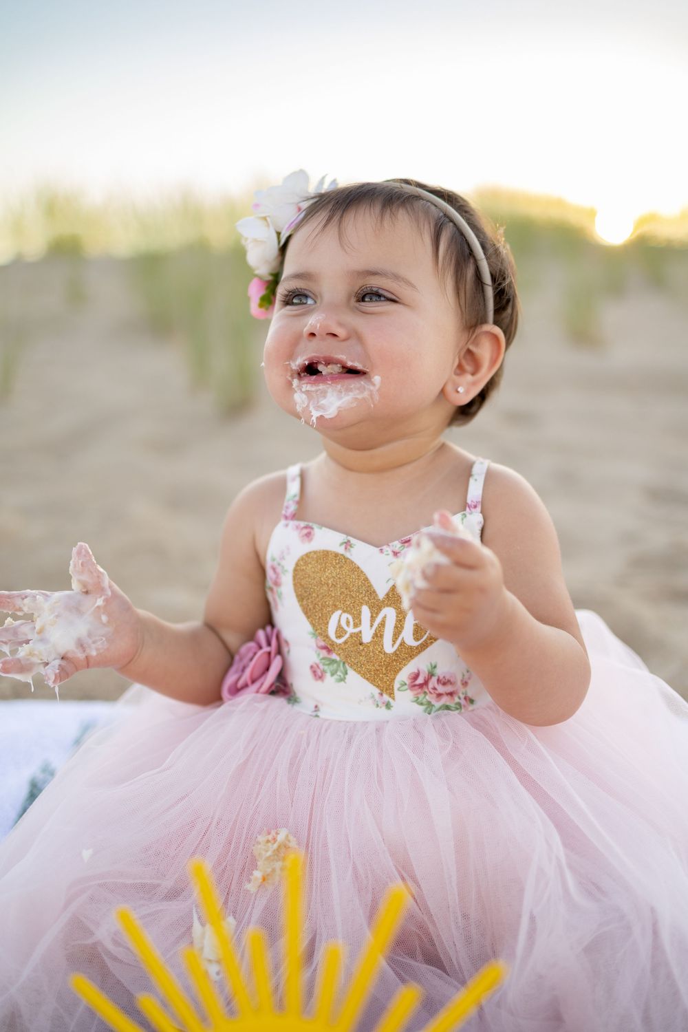 A baby girl in a pink tutu dress celebrating her first birthday with cake on her face at a beach.