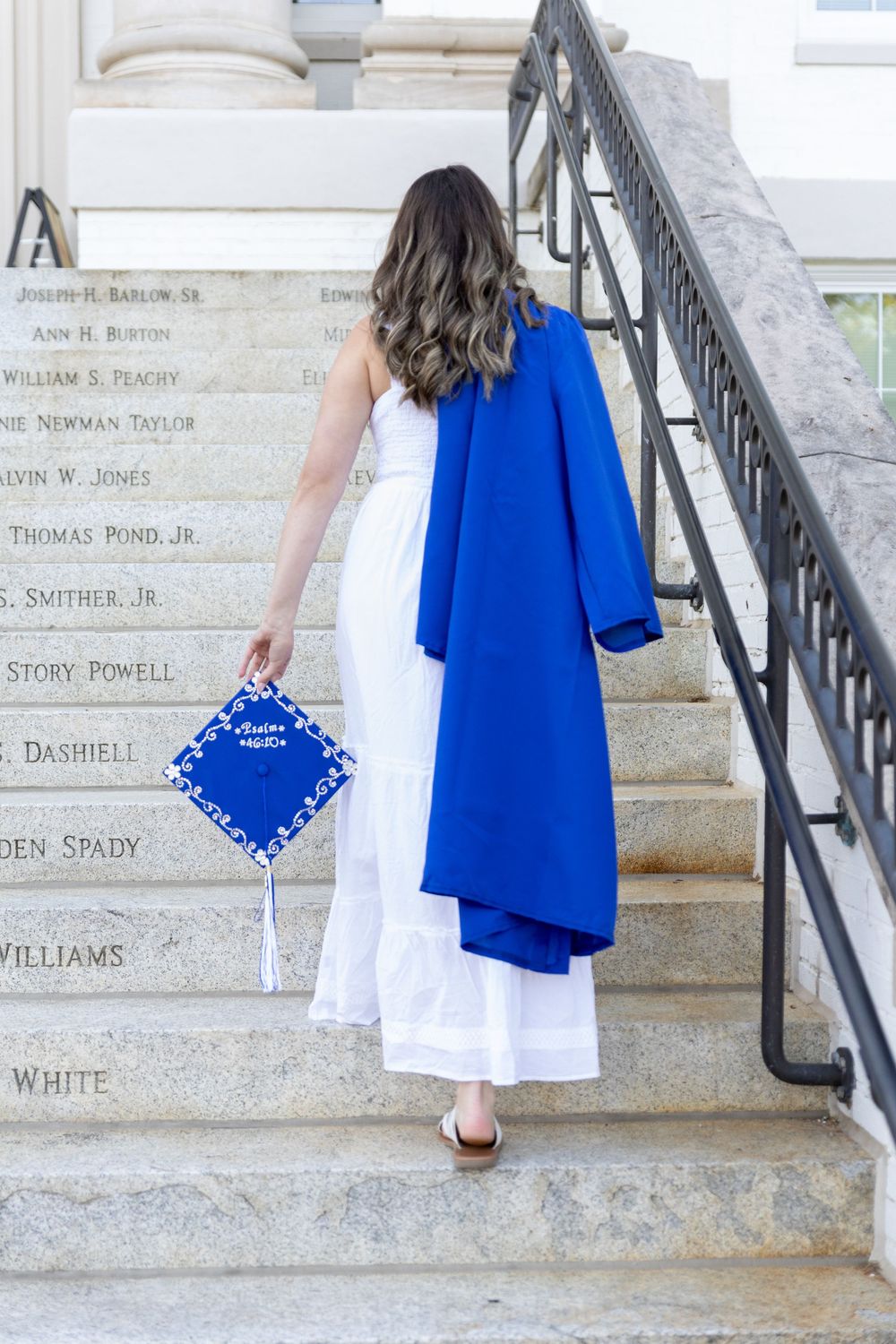 A graduate walks up stone steps in a white dress, holding a blue cap and gown, with names engraved on the staircase.