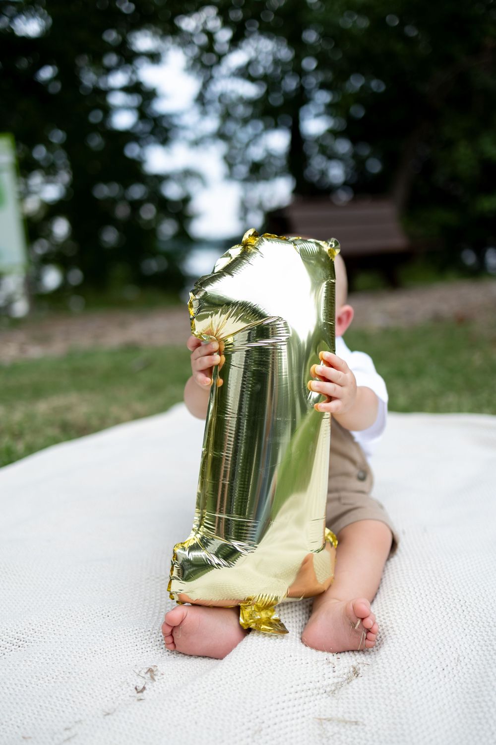 Baby sitting outdoors holding a gold number one balloon on a white blanket, celebrating their first birthday.