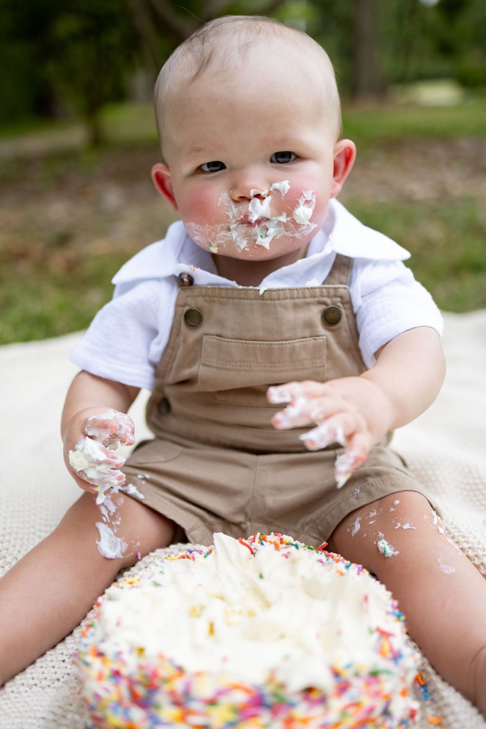 Baby boy in overalls enjoying his first birthday cake smash outdoors, with frosting and sprinkles on his face and hands.