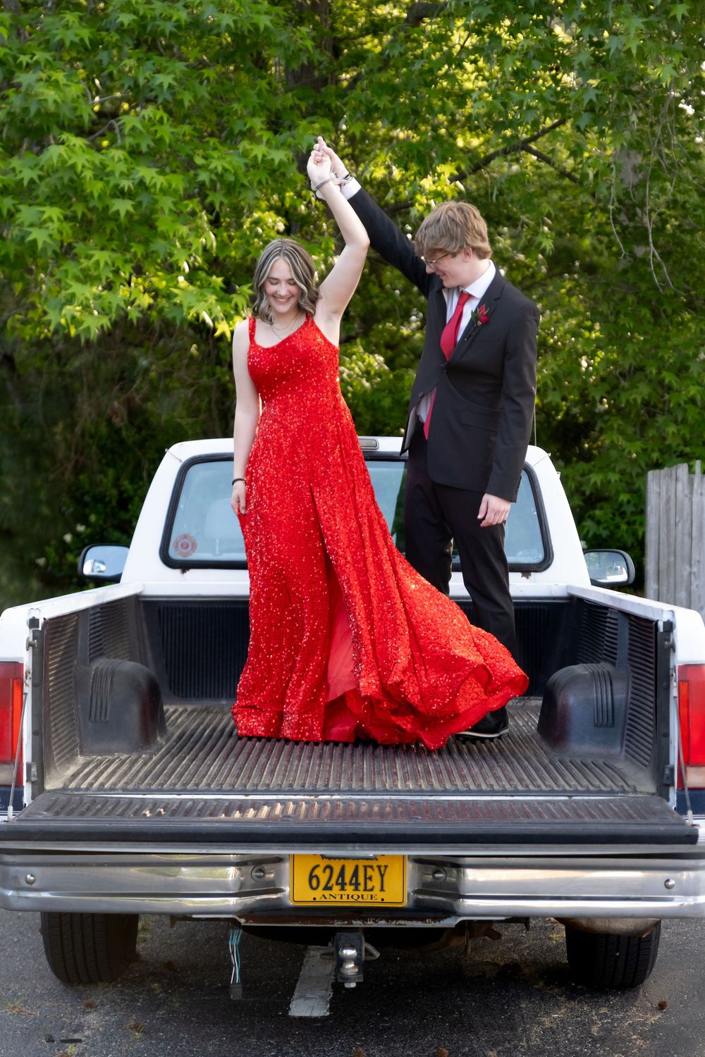 Teen couple in prom attire dancing on pickup truck bed, girl in red gown, boy in suit, green trees background.