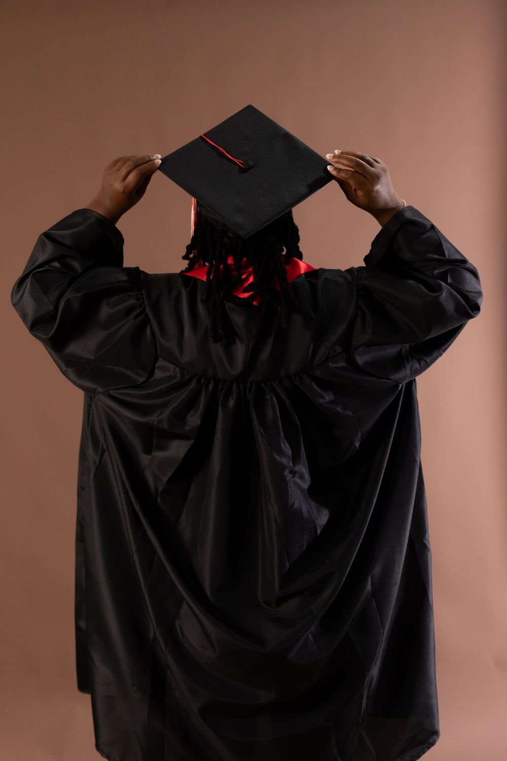 Graduate in black cap and gown seen from behind, adjusting mortarboard with red tassel on brown background.