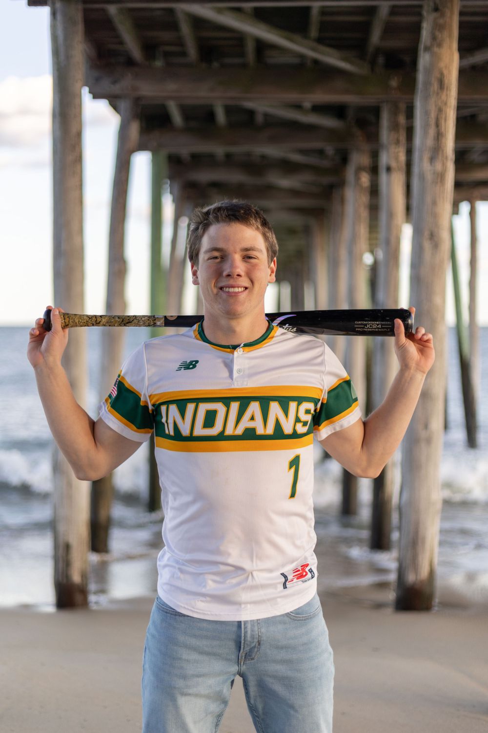 Baseball player in Indians jersey poses with bat under pier at beach.