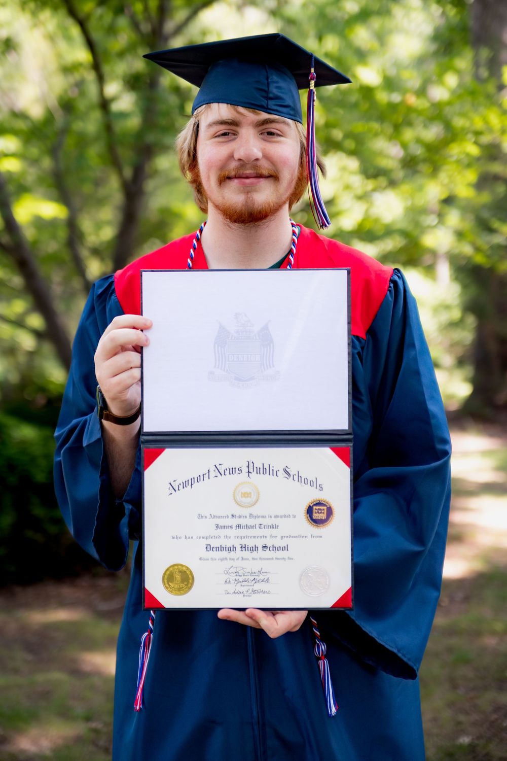A Denbigh graduate in cap and gown holds up their diploma from Newport News Public Schools while standing outdoors.