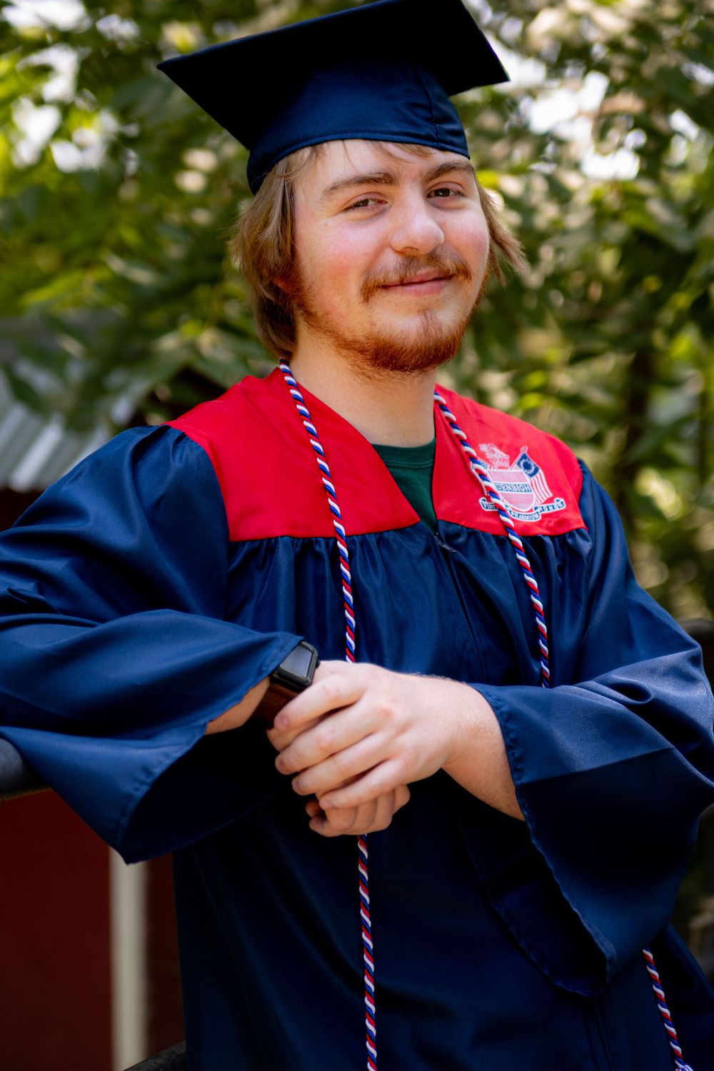 Denbigh High School Graduate in navy cap and gown with red stole smiles while standing outdoors on graduation day.