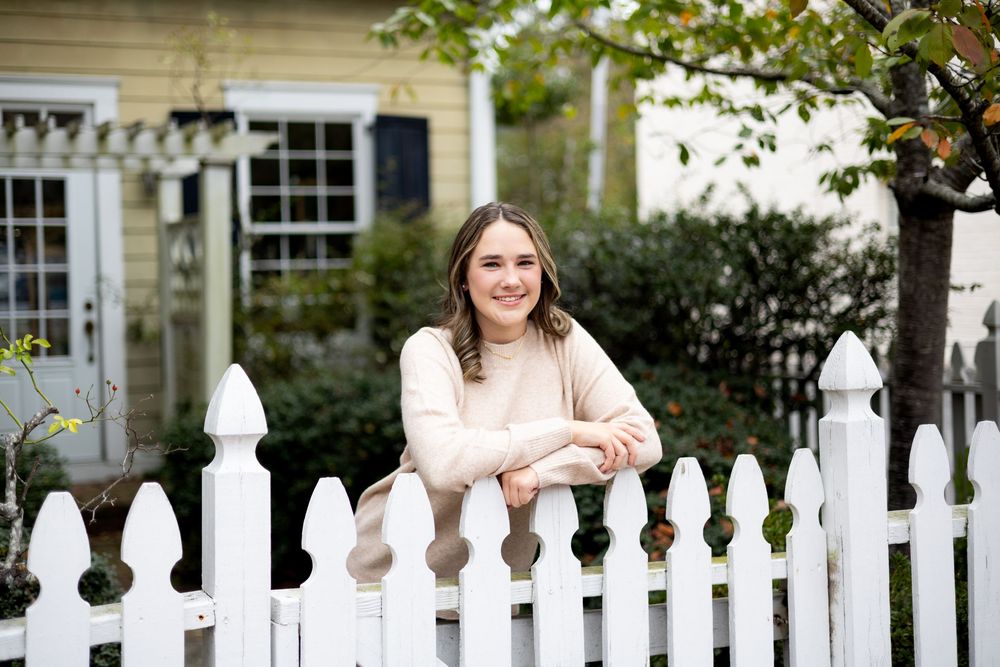 People gather behind a white picket fence in front of a traditional home with windows and landscaping.