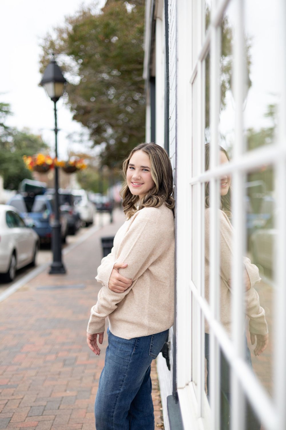 Person in beige sweater and jeans poses against white building exterior with brick sidewalk.