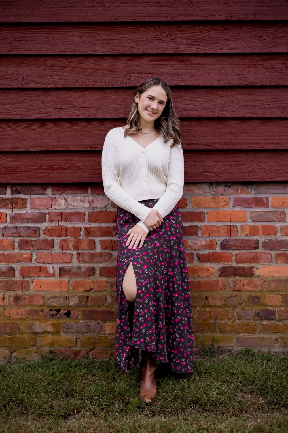 Woman in white sweater and floral skirt sits casually against rustic red barn wall and brick foundation.
