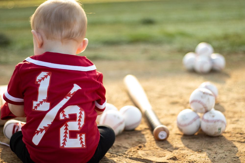 A young baseball fan in a red jersey sits with baseball equipment on a field.