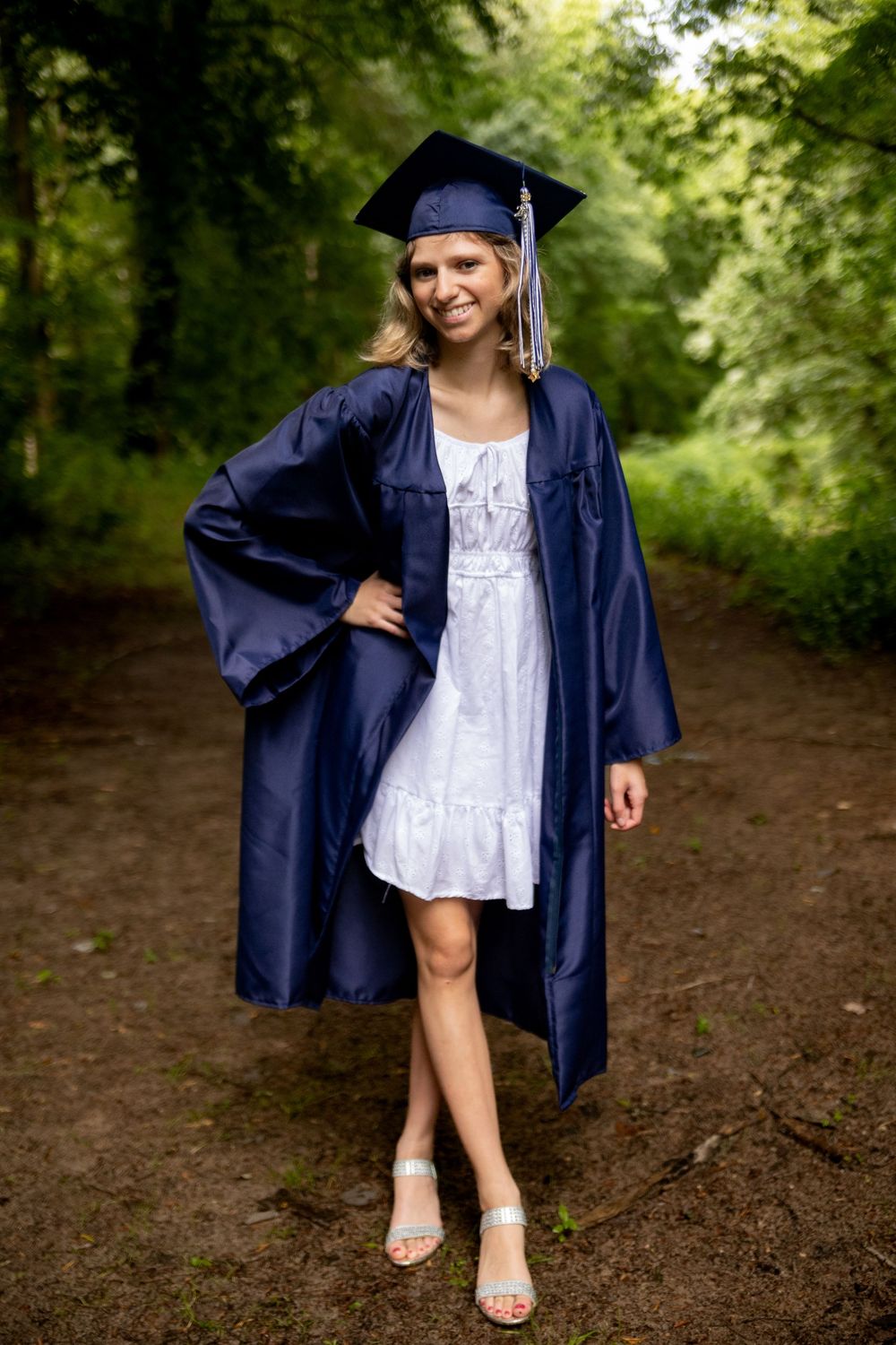 A graduation photo in a navy blue cap and gown with a white dress underneath, taken on a wooded path.