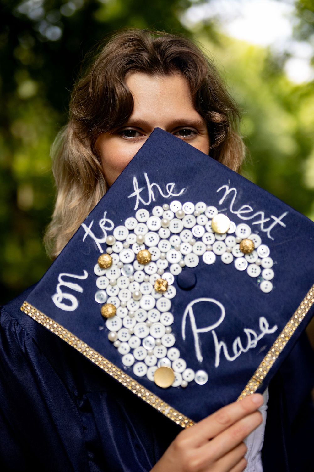 A decorated graduation cap features 'On to the next Phase' written with pearls and gold accents on navy blue fabric.