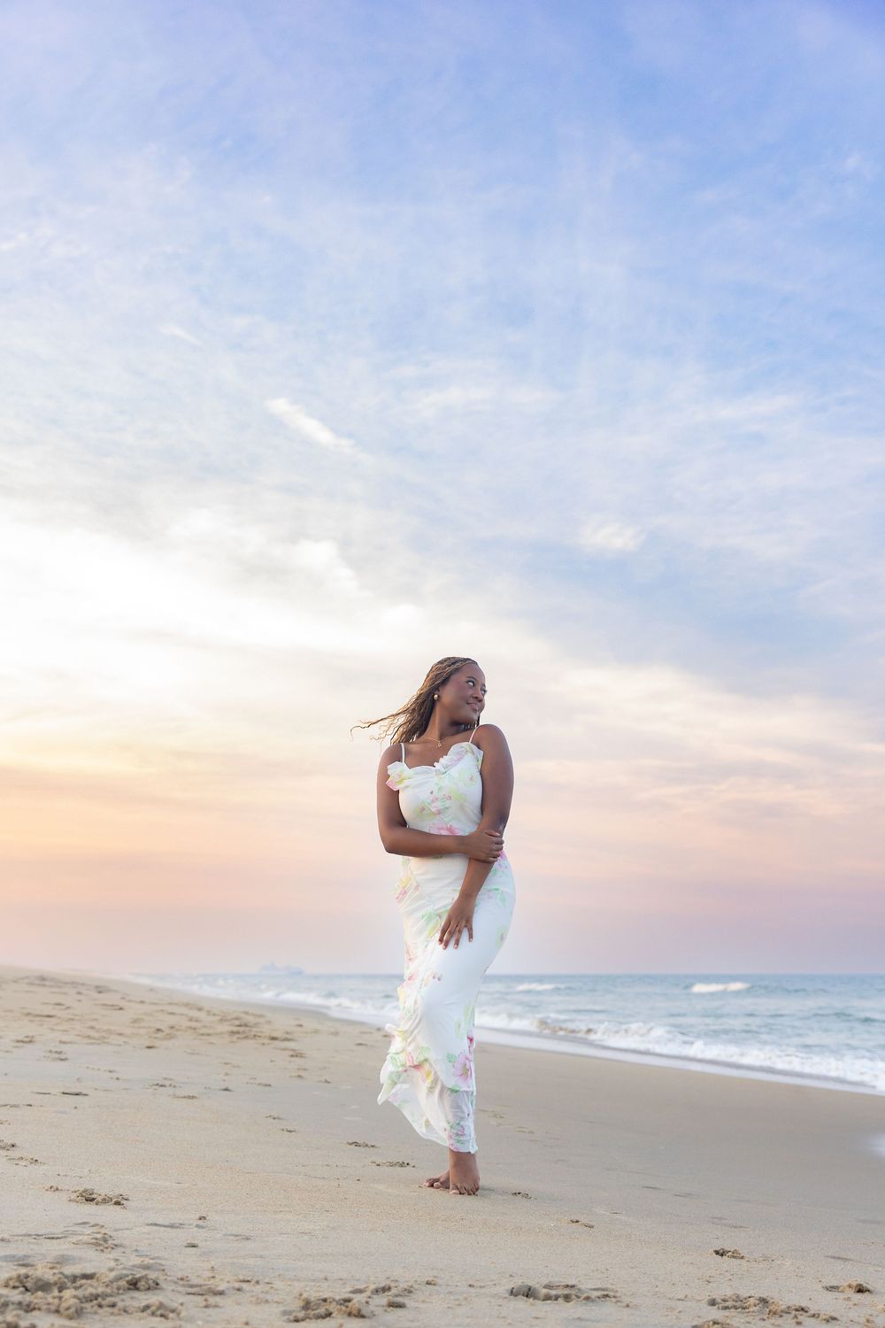 Woman in flowing white dress dances gracefully on sandy beach at sunset with ocean waves in background.