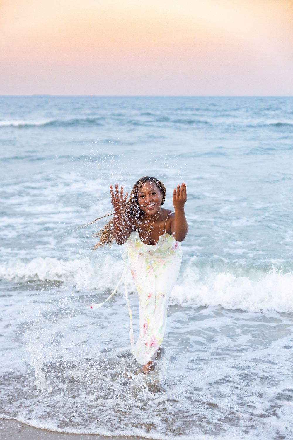 A person in a white dress splashes joyfully in ocean waves during sunset with water spraying around them.