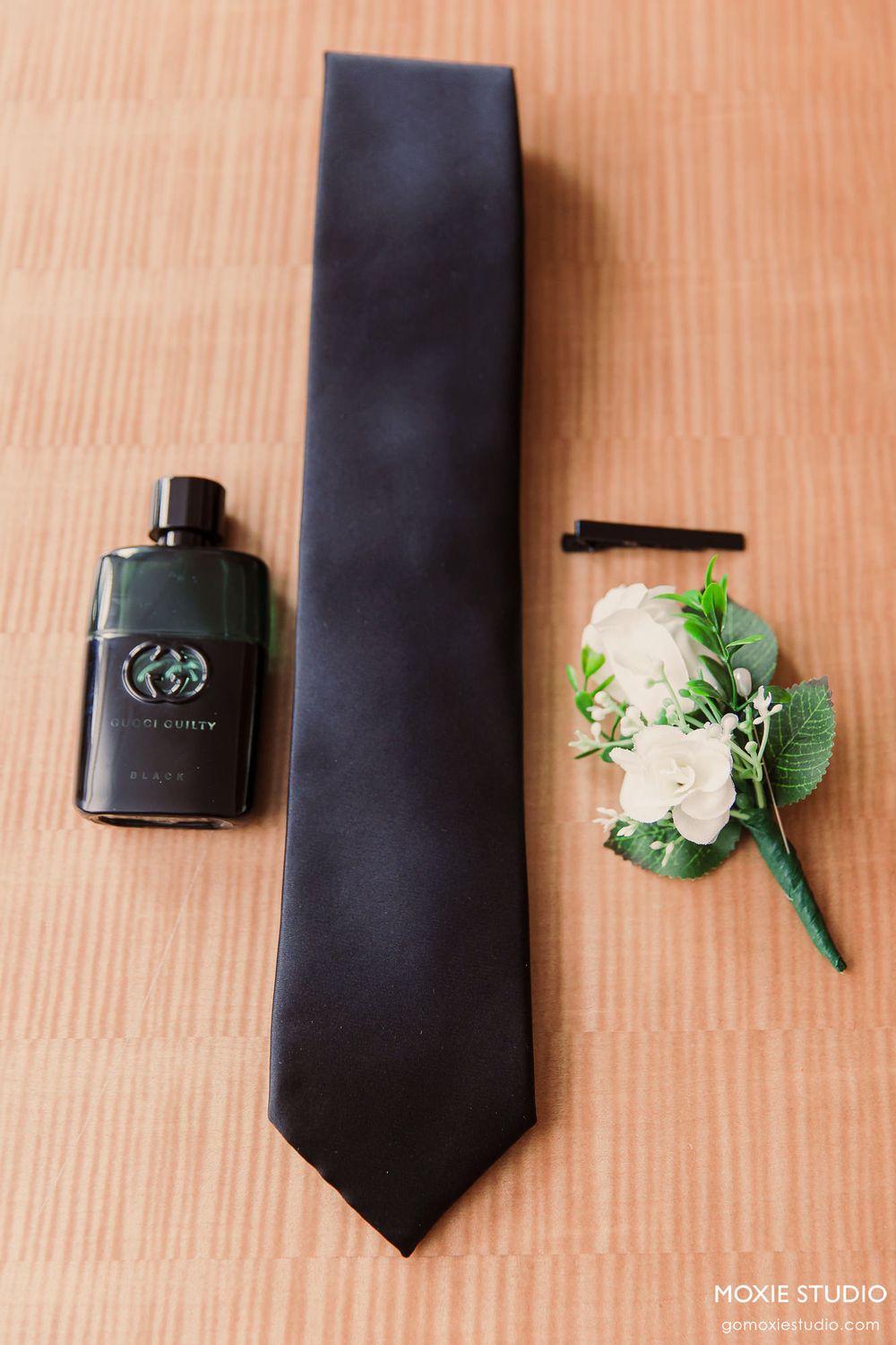 Black silk necktie laid flat with white floral boutonniere and cologne bottle arranged on wooden surface.