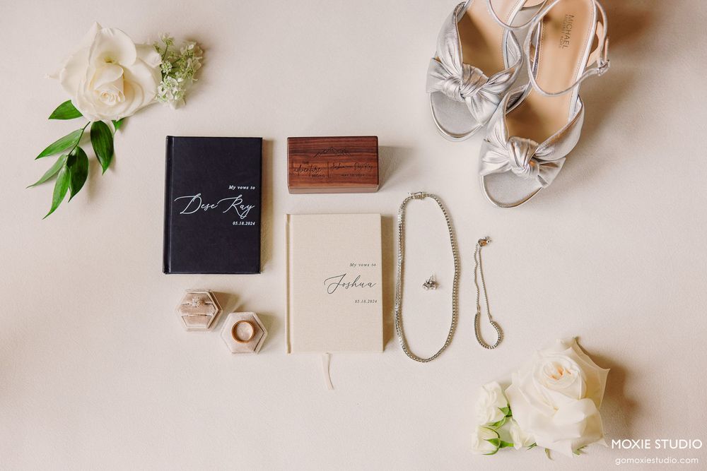 Wedding details including invitations, rings, shoes, and floral elements arranged on a light beige surface.