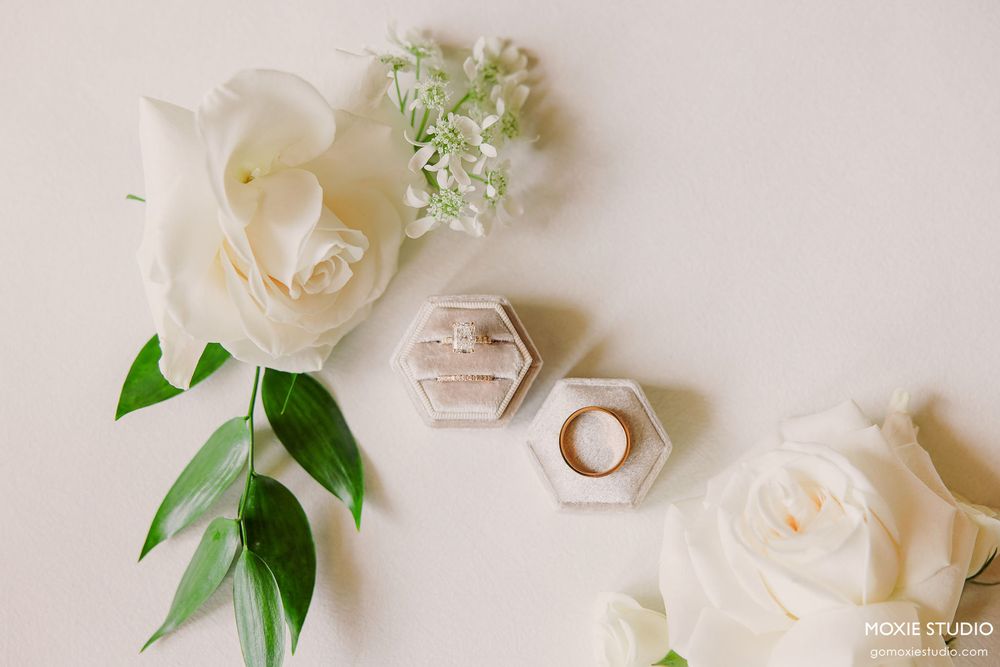 Wedding rings and delicate white flowers arranged on a light beige surface with green foliage accents.