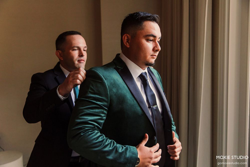 Groomsmen in black suits and green vests stand in a formal line while adjusting their ties and cuffs.