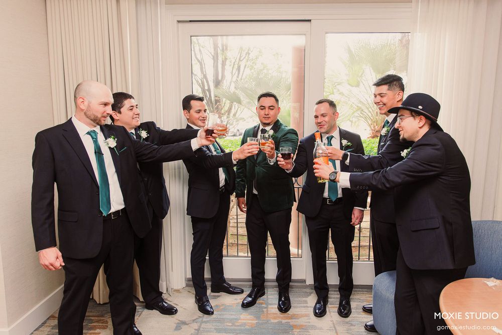 Groomsmen in black suits line up against a beige wall in a wedding venue hallway.