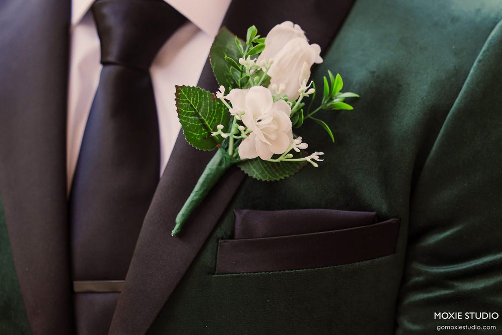 A white rose and greenery boutonniere pinned to a dark suit lapel with delicate floral details.
