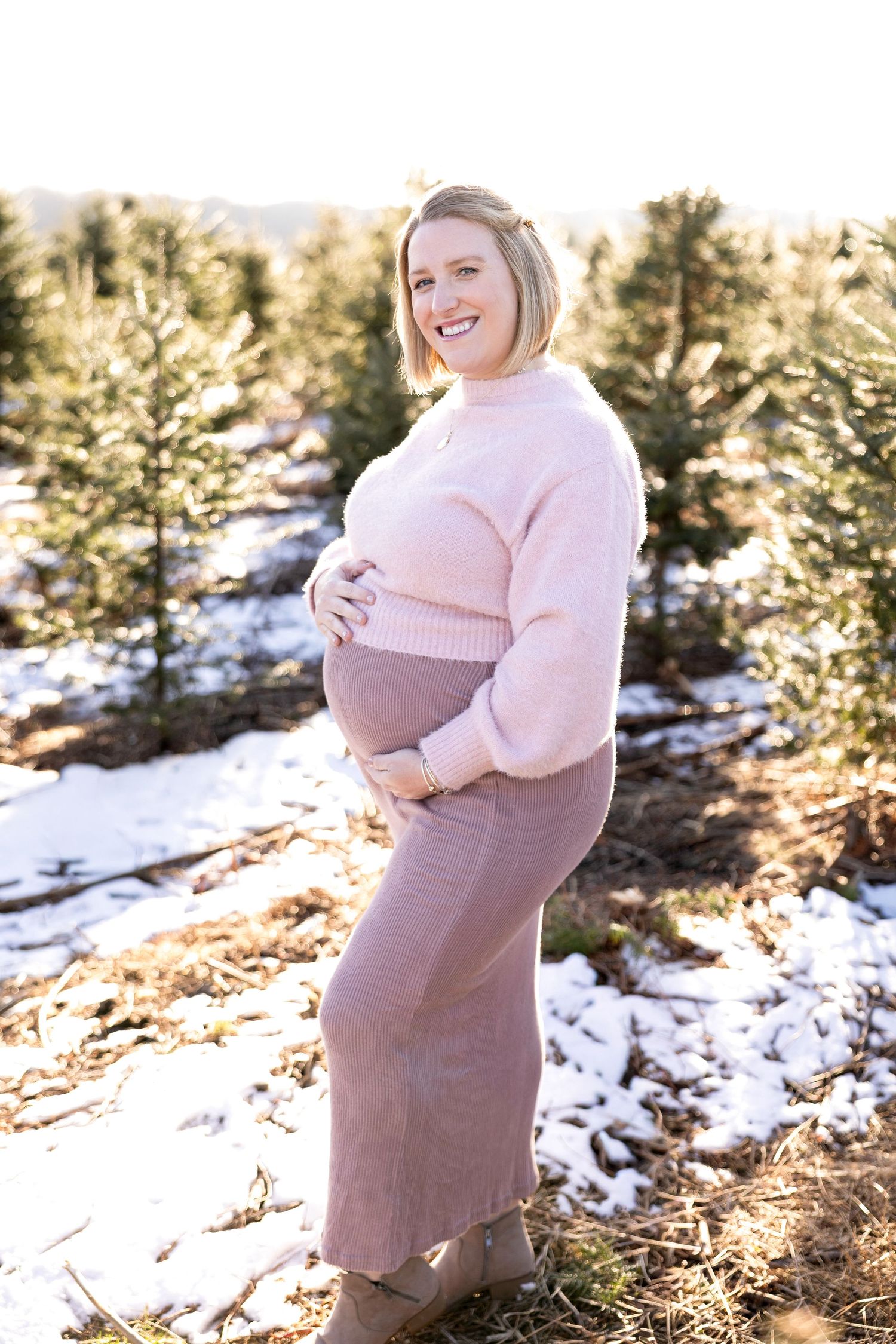 Winter maternity photoshoot in snowy forest with pink sweater.