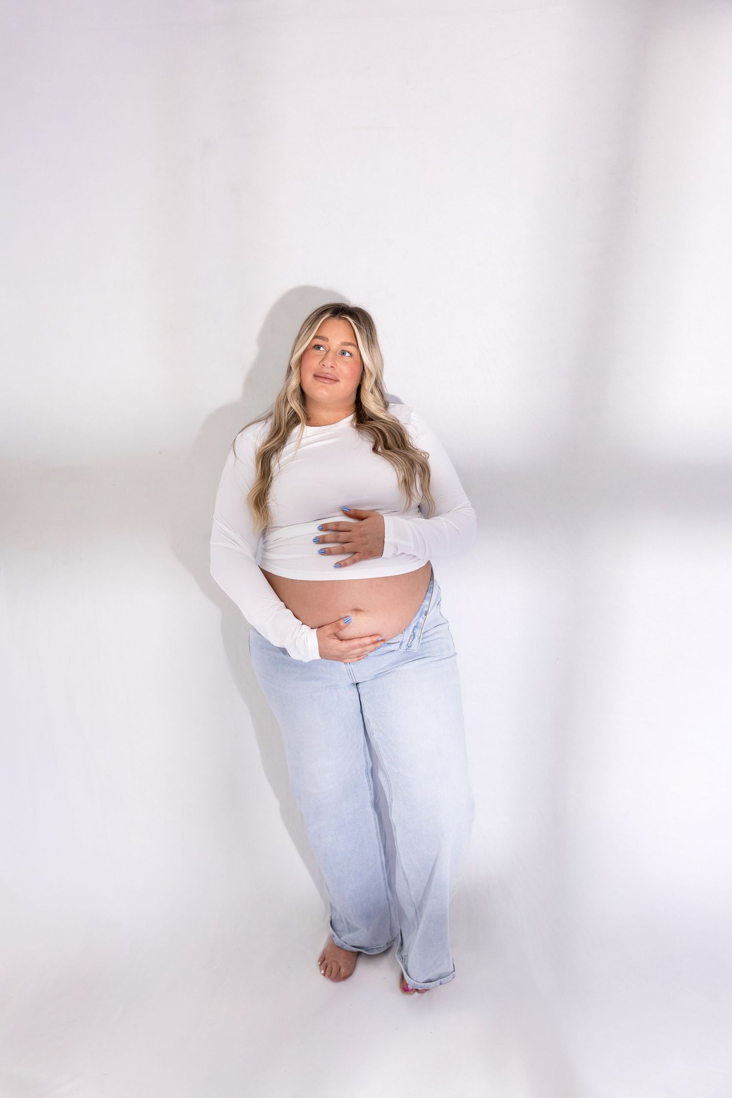 Maternity photos of woman in light denim and white crop top against white background.