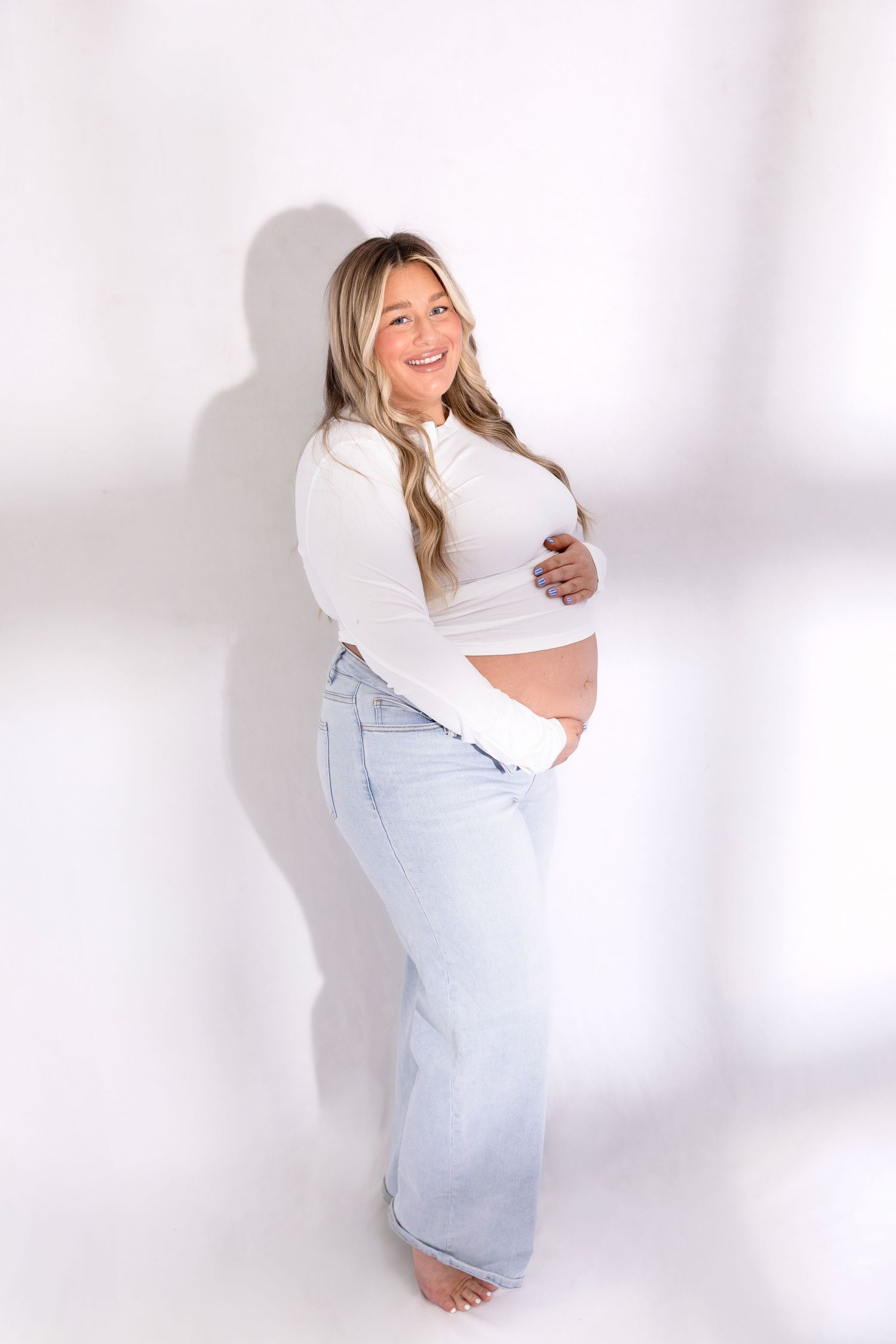 Maternity photo shoot in white top and light jeans against white background showing growing baby bump.