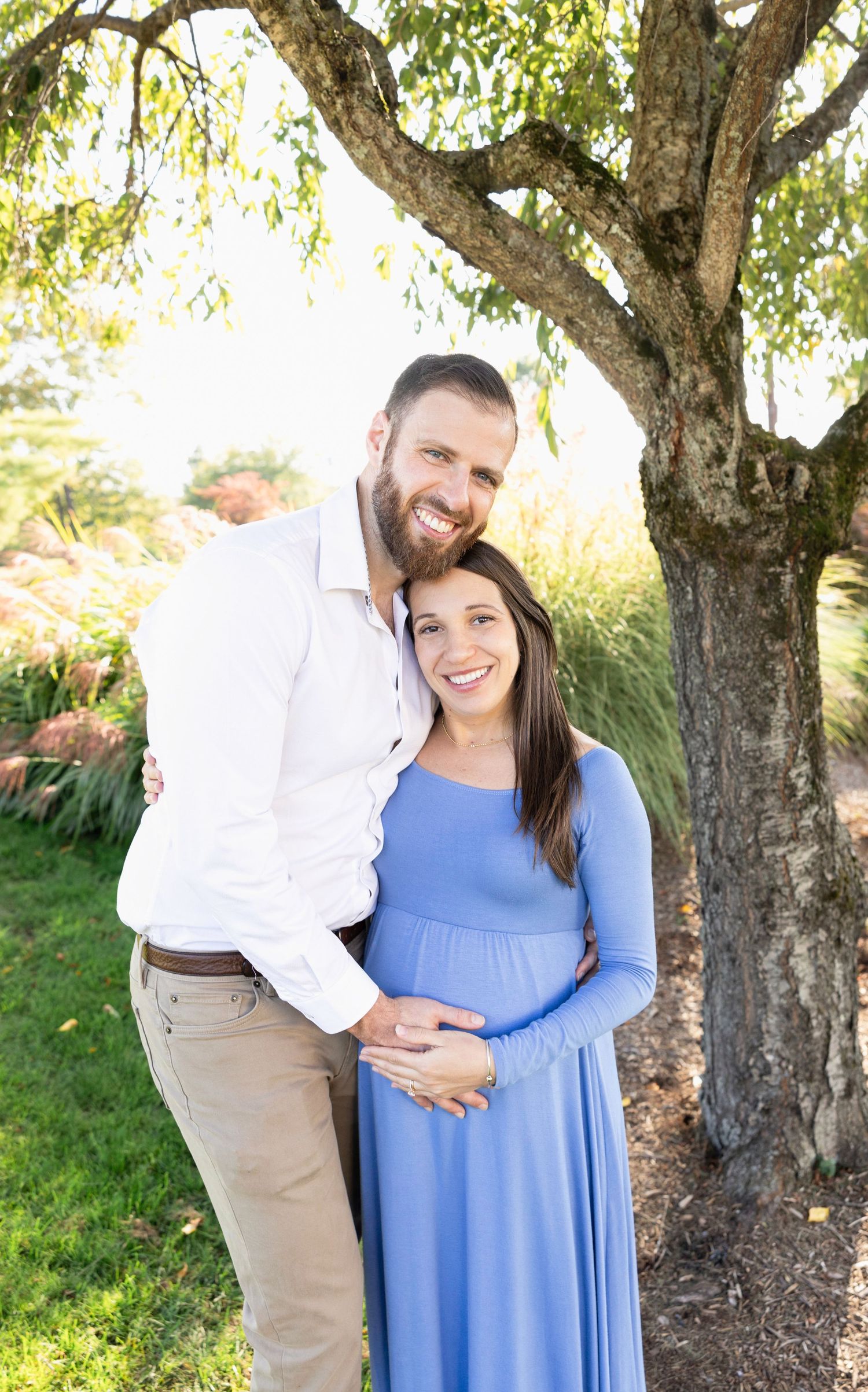 Couple poses under tree branches in outdoor maternity photoshoot with blue dress.