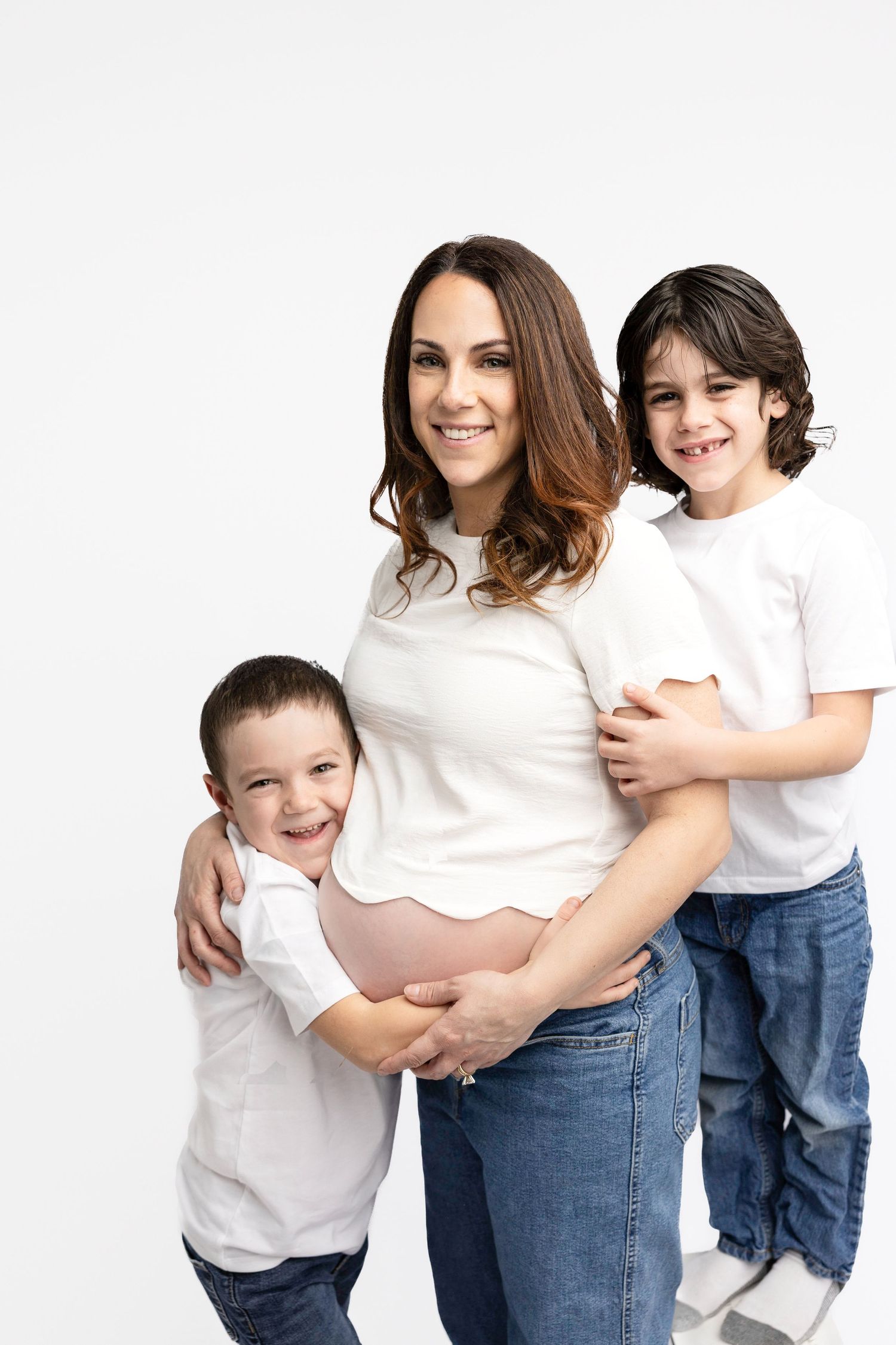 Maternity family photos with children in matching white shirts and jeans against white backdrop.