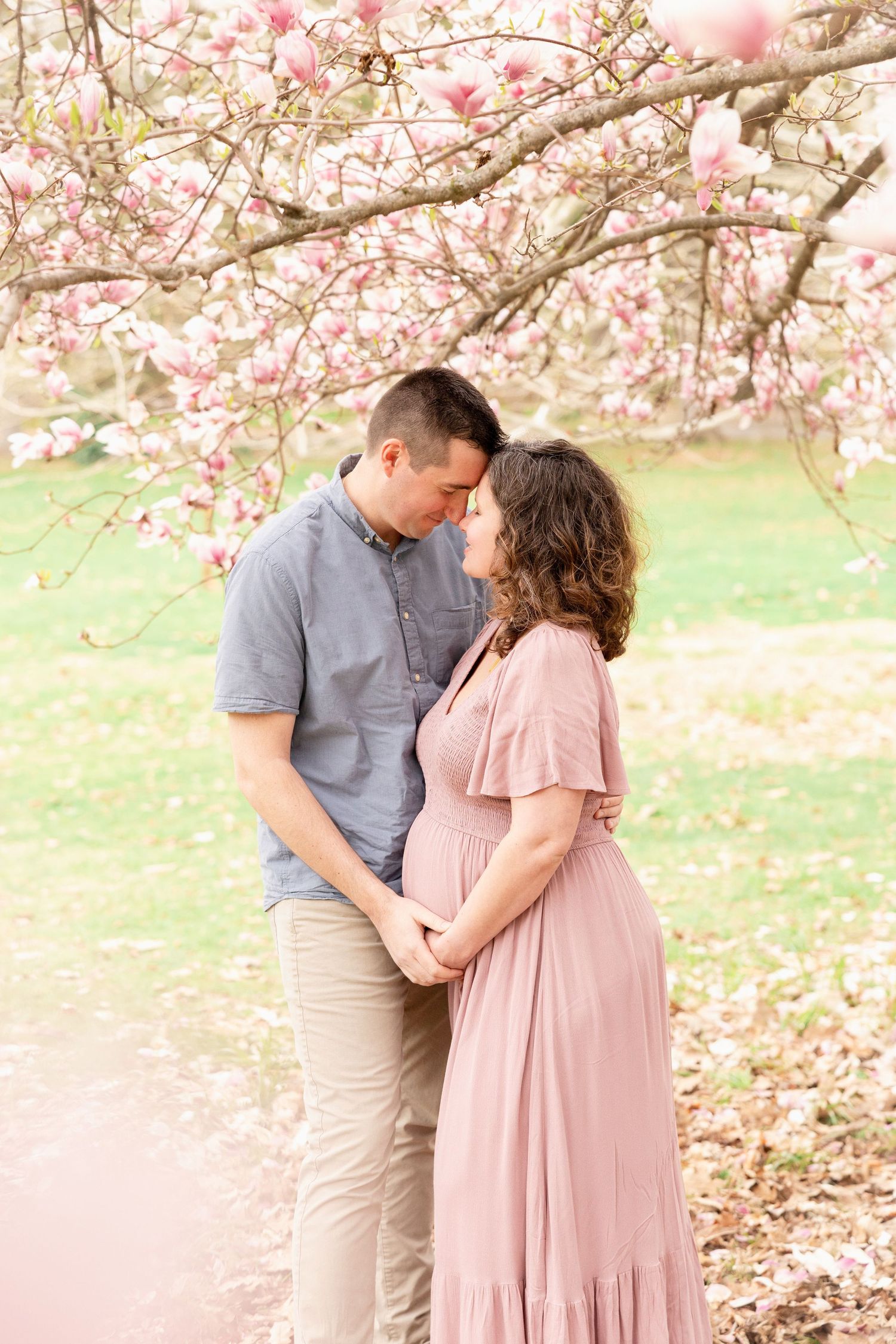 Couple sharing a tender moment under blooming pink magnolia tree, pregnant woman in mauve dress holding hands with partner.