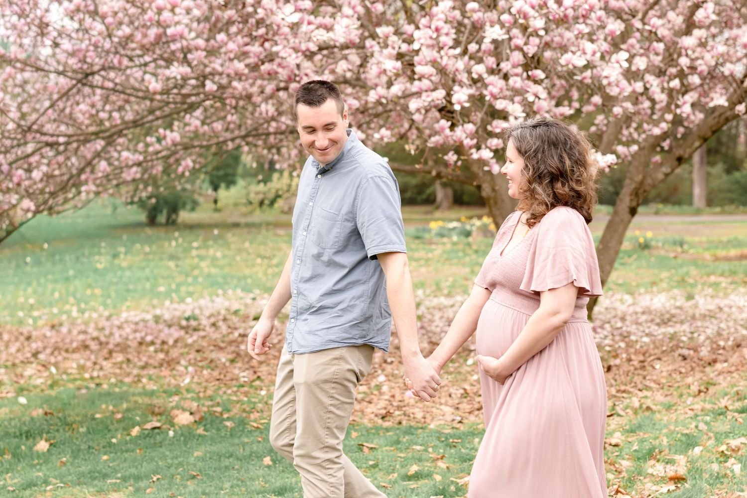 Couple holding hands under blooming pink cherry blossom tree, woman in pink maternity dress, spring outdoor maternity photo.