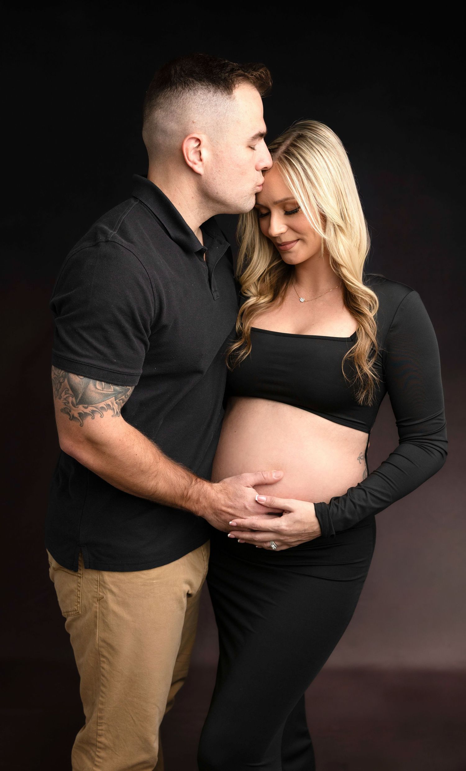 Intimate maternity photo session with couple in black and khaki against dark background.