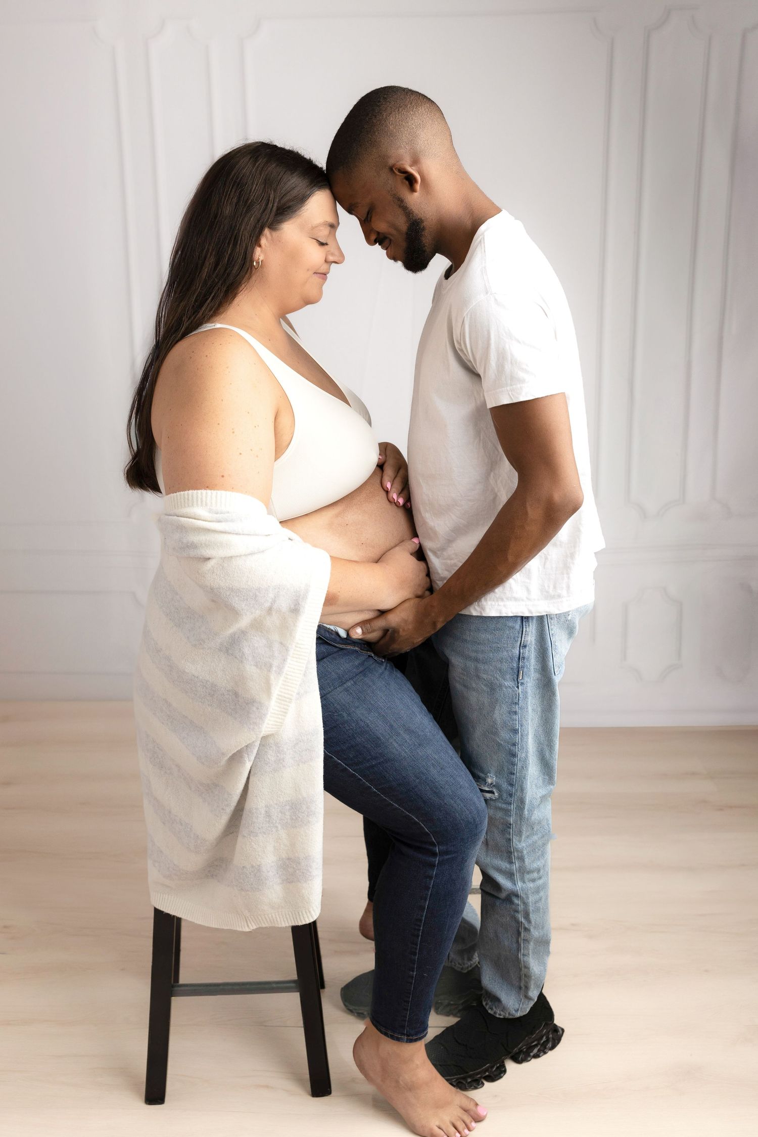 Intimate maternity photoshoot with couple in white shirt and jeans against white wall.