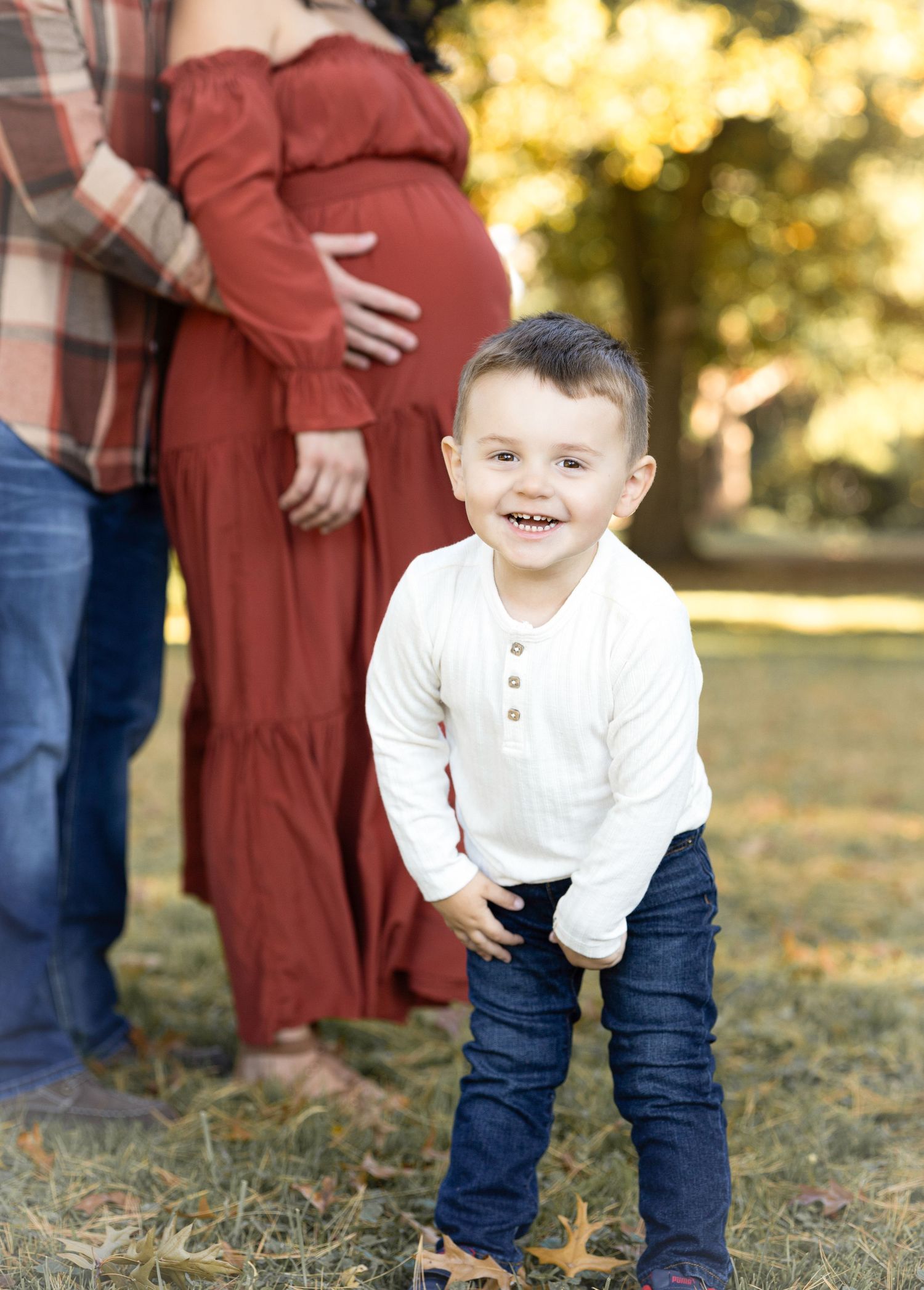 A small child in white and denim plays in fallen autumn leaves during an outdoor photo session.