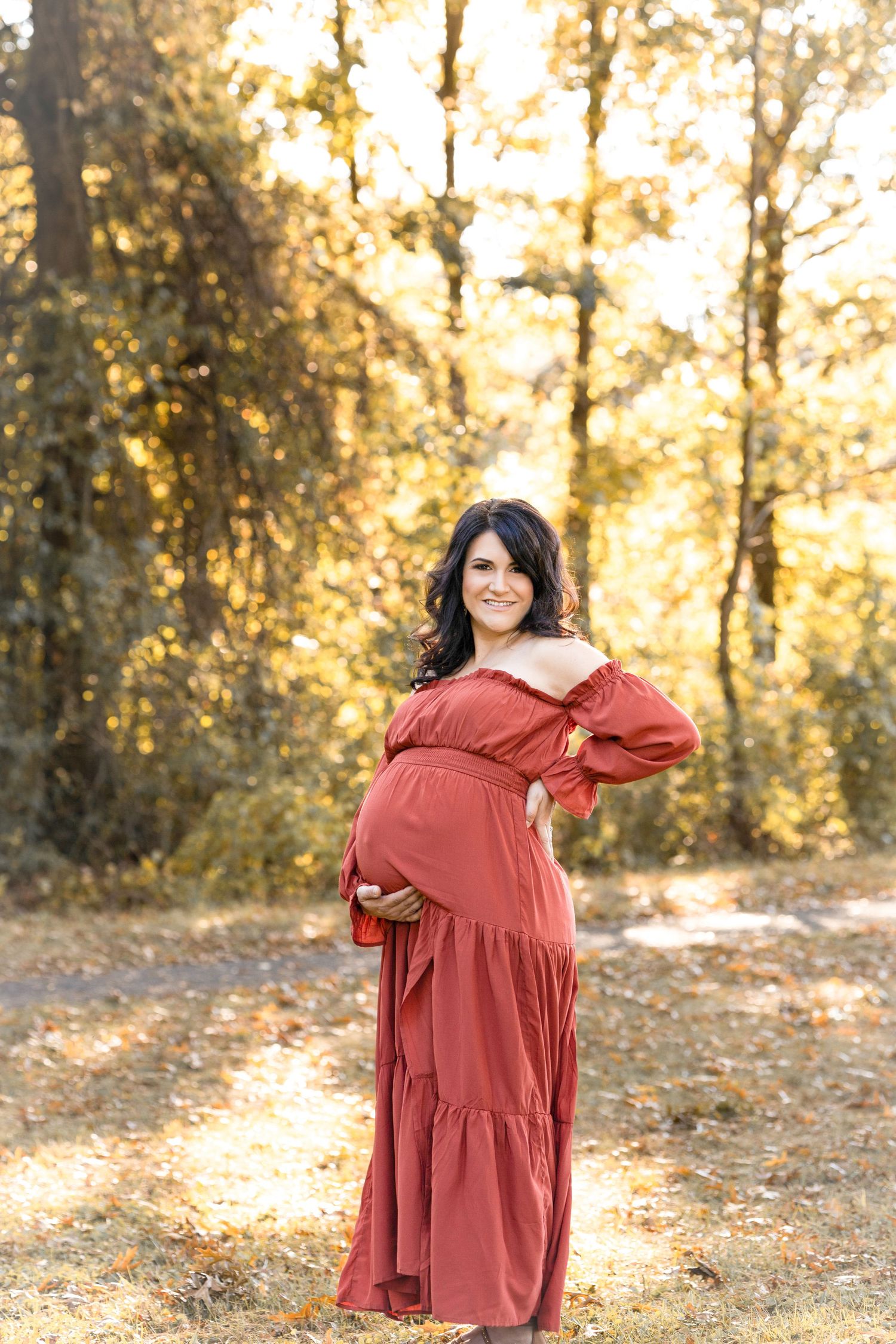 A person in a rust-colored dress poses playfully in a wooded area during golden hour.