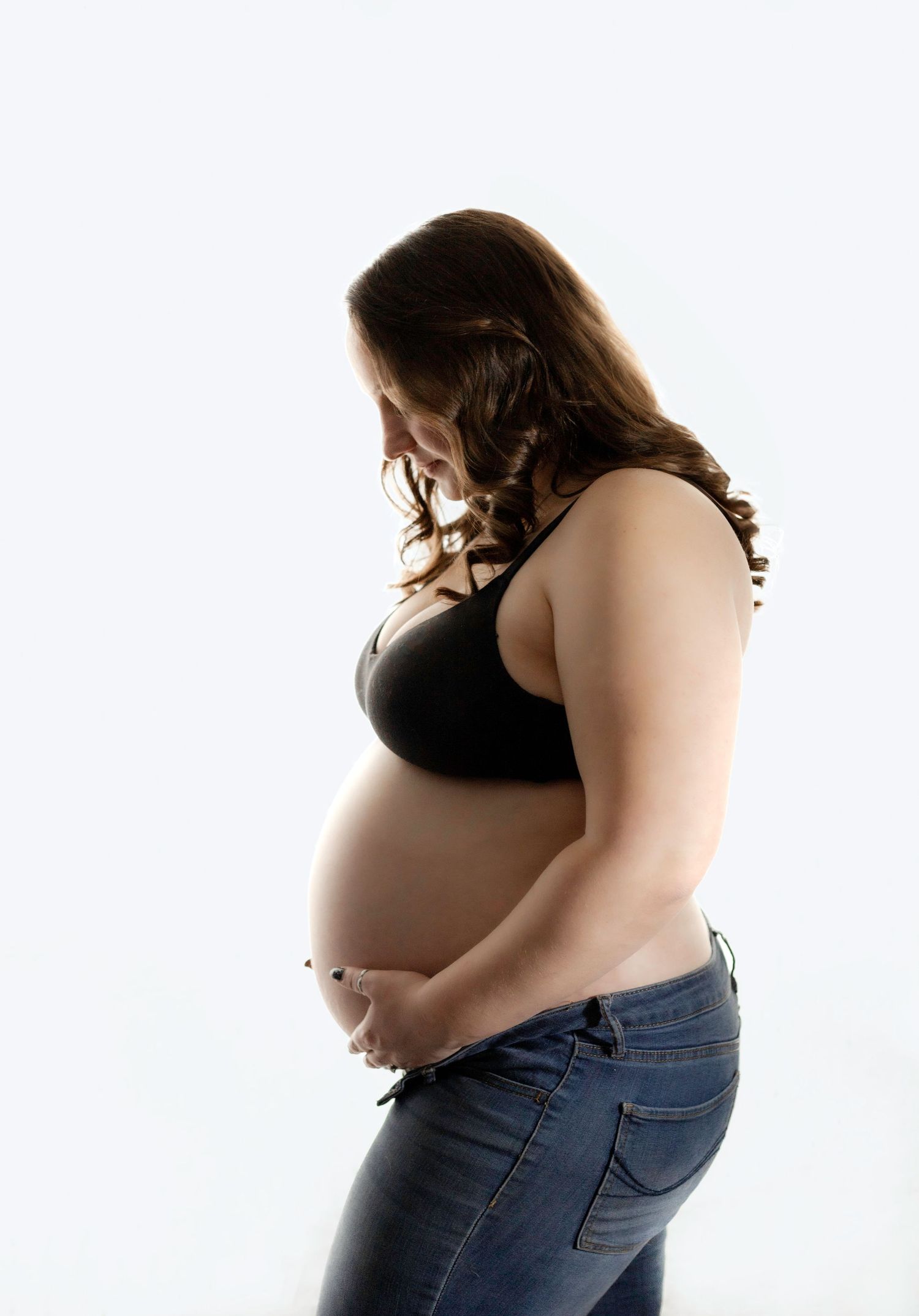 Side profile of pregnant woman in black top and jeans against white background.