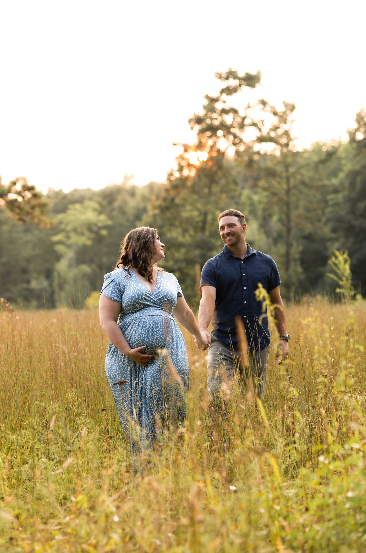Romantic couple walks through tall grass field during golden hour photoshoot.
