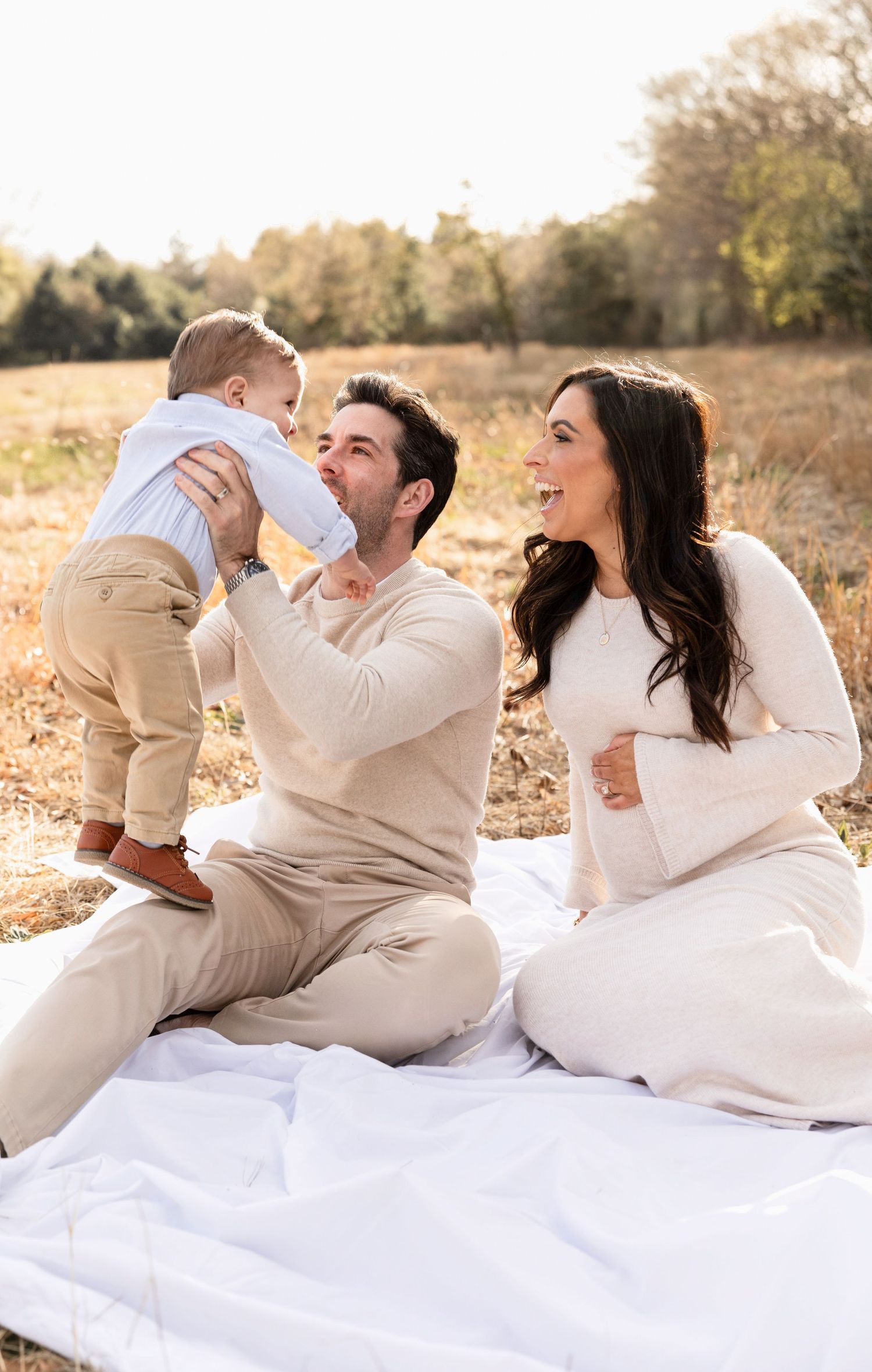 Family in cream colored outfits lounging on white blanket during outdoor photoshoot at sunset.