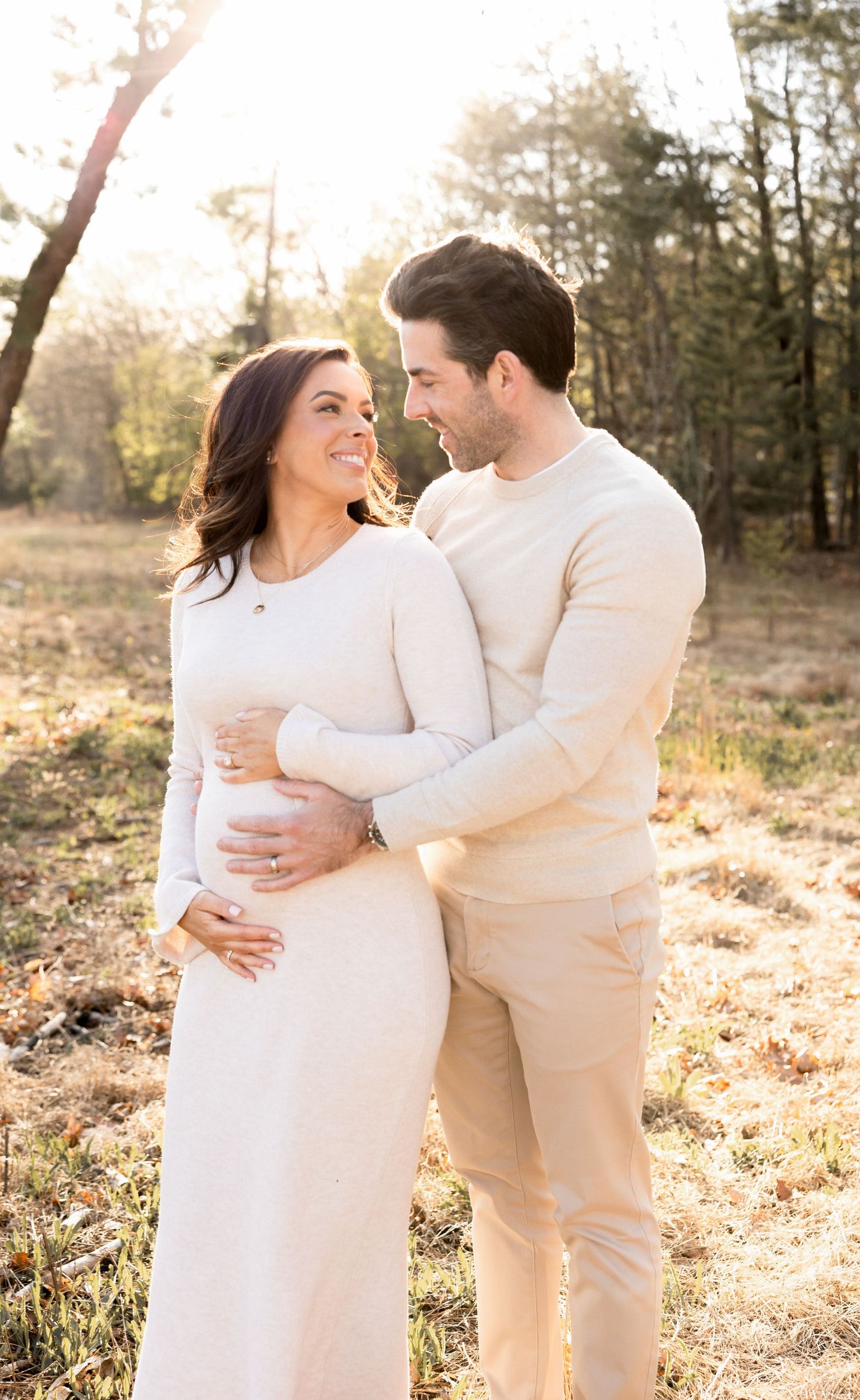 Couple in matching cream outfits embrace during sunset maternity photoshoot in nature.