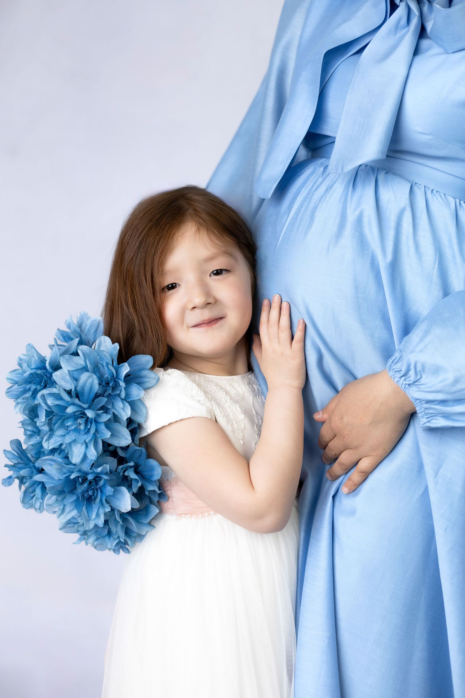 Child in white dress poses with person wearing light blue outfit holding blue flowers.