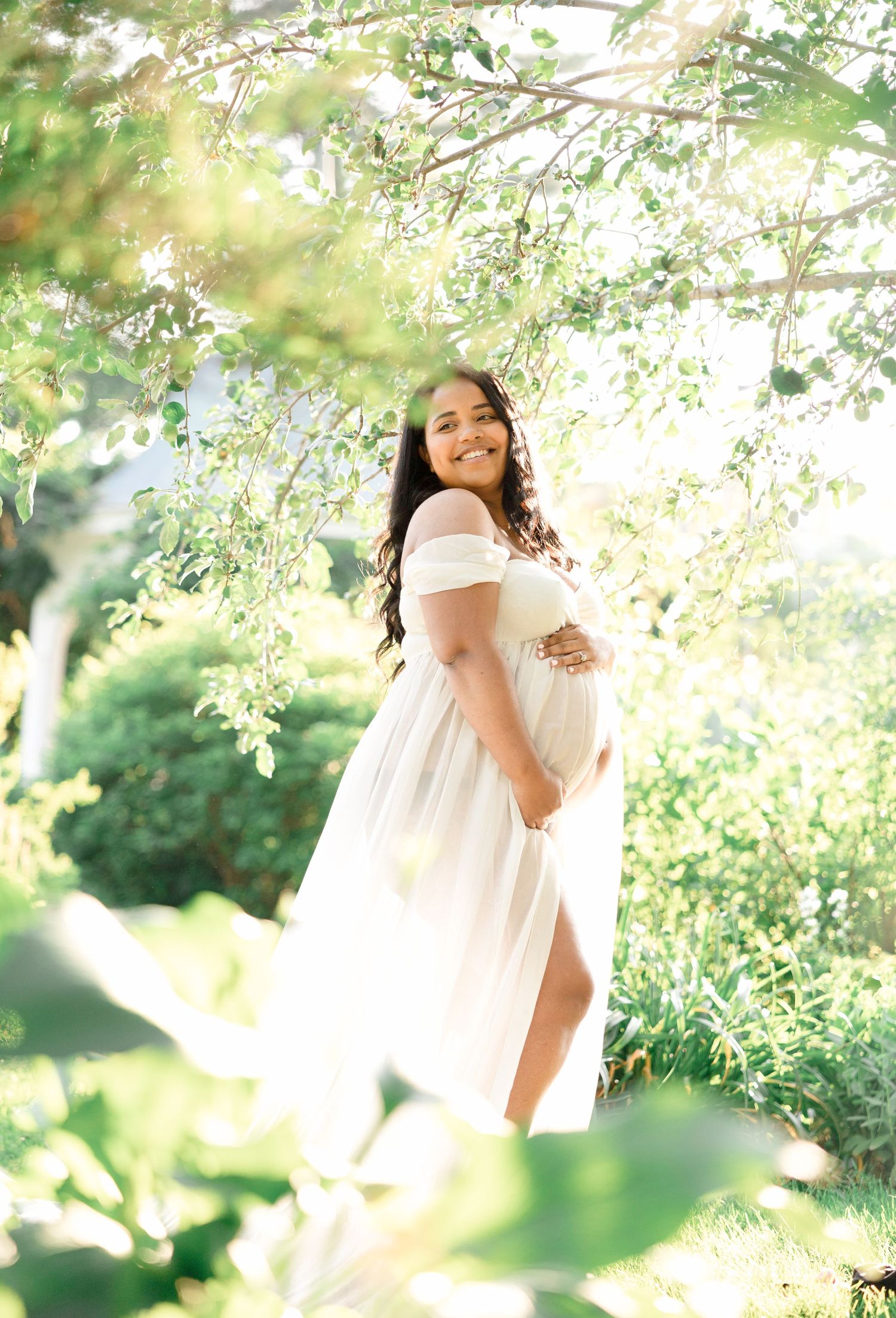 Maternity photo of woman in flowing white dress under blooming tree branches.