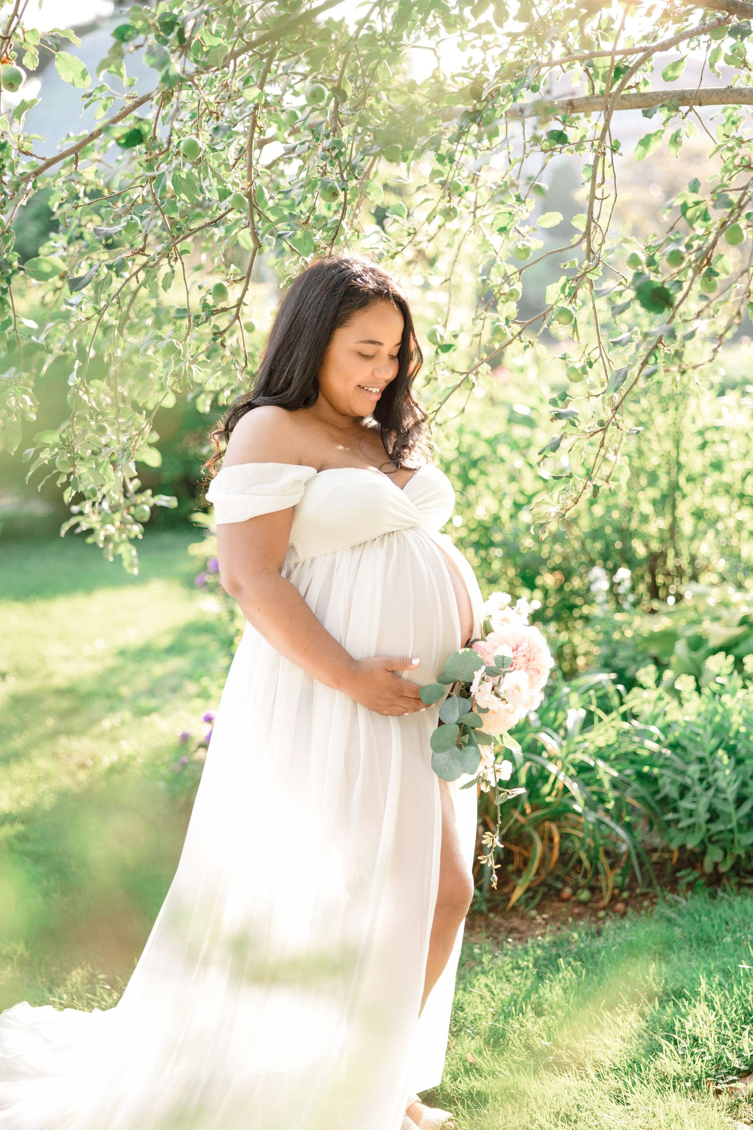 Ethereal maternity portrait with woman in white dress holding flowers under tree branches.