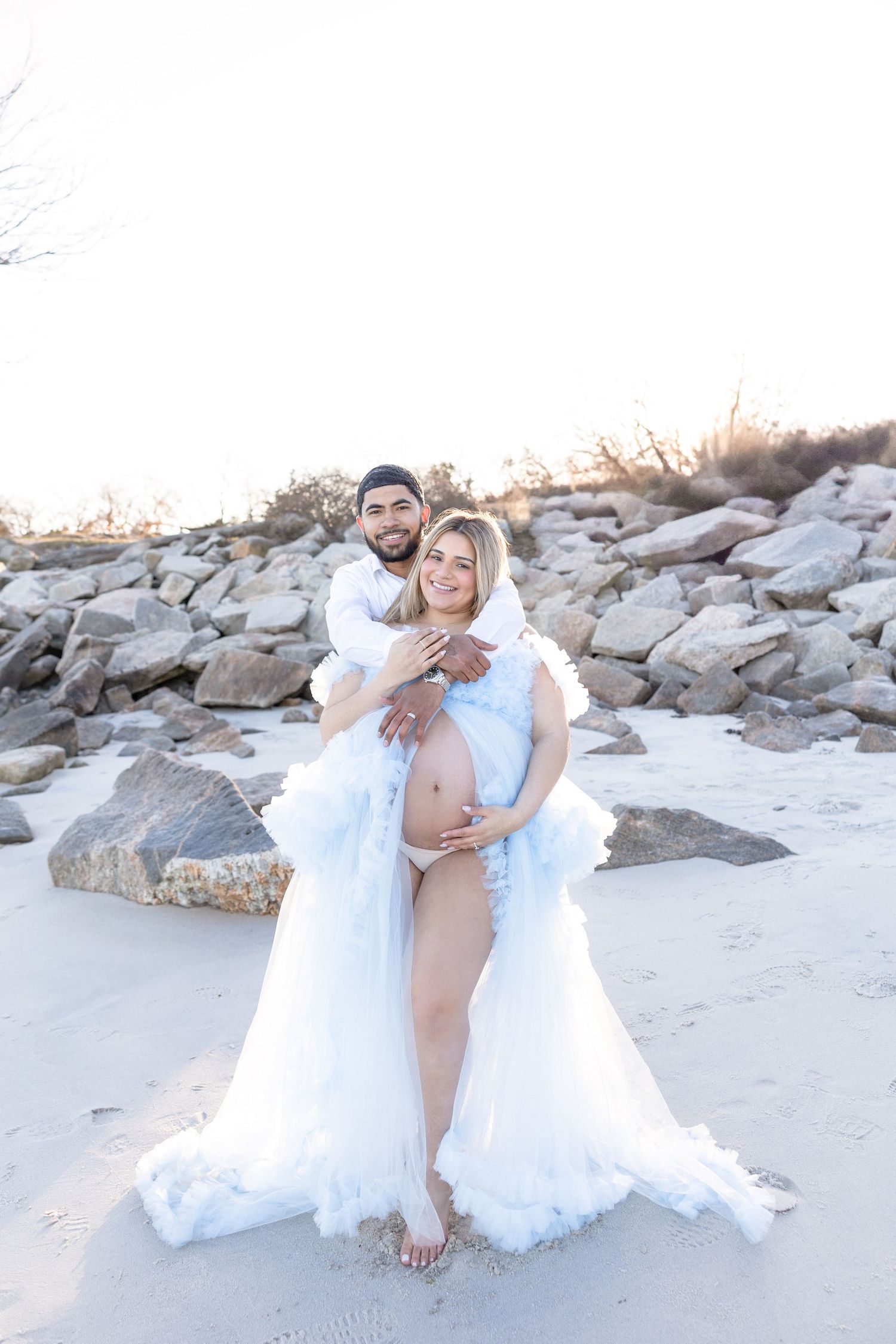 Couple in white outfits embraces on beach with rocky backdrop at sunset.