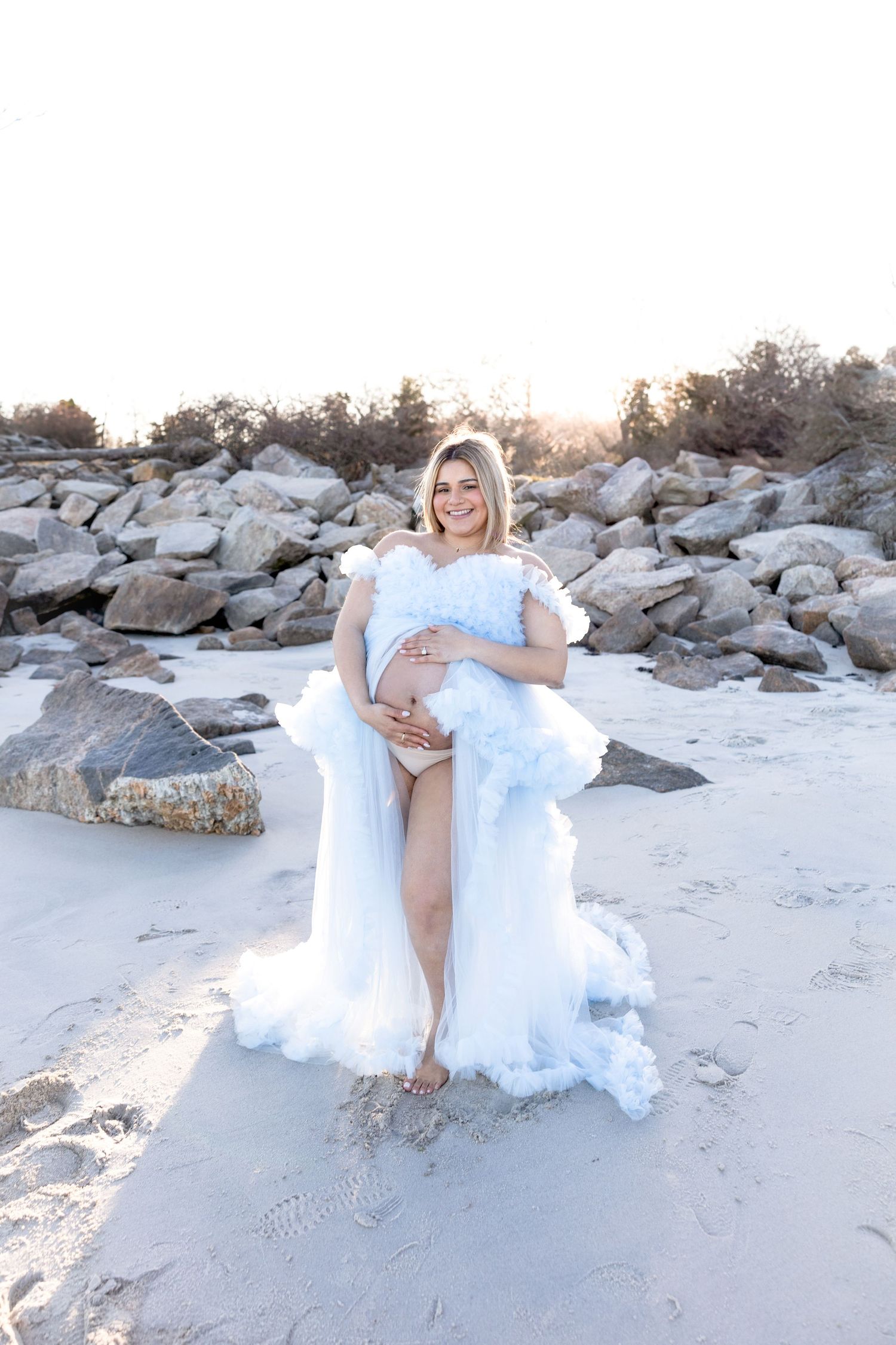 Maternity photos on beach with flowing light blue dress against rocky landscape.