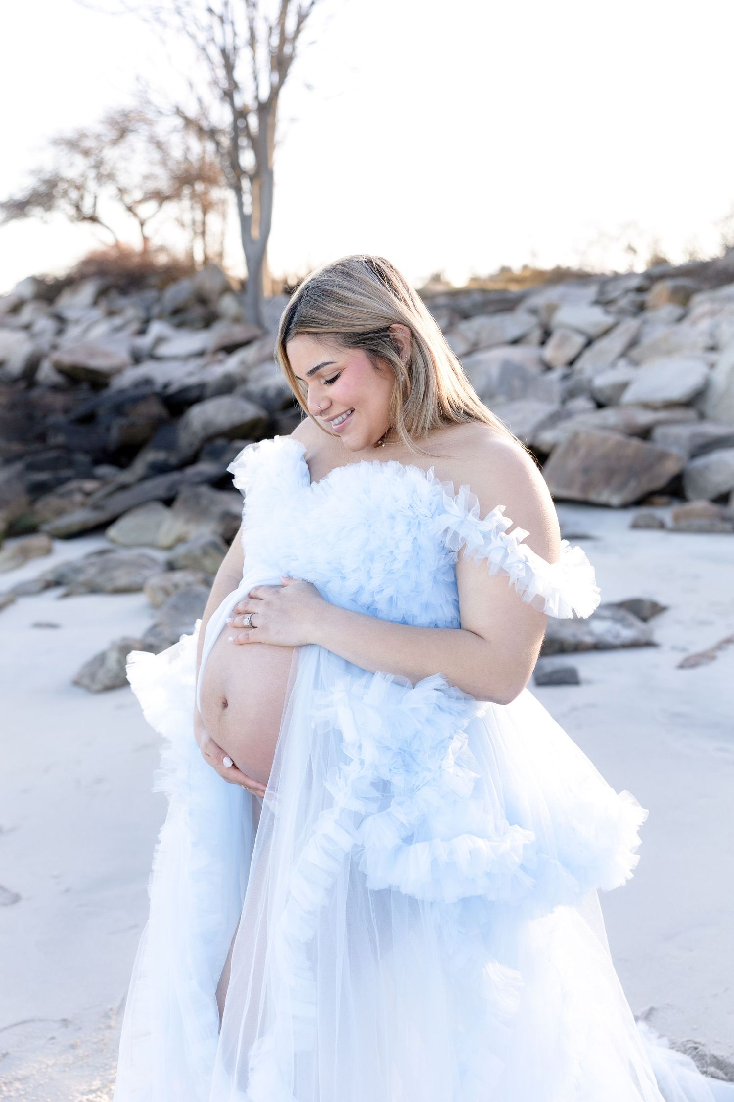 Maternity session features flowing light blue dress on sandy beach.