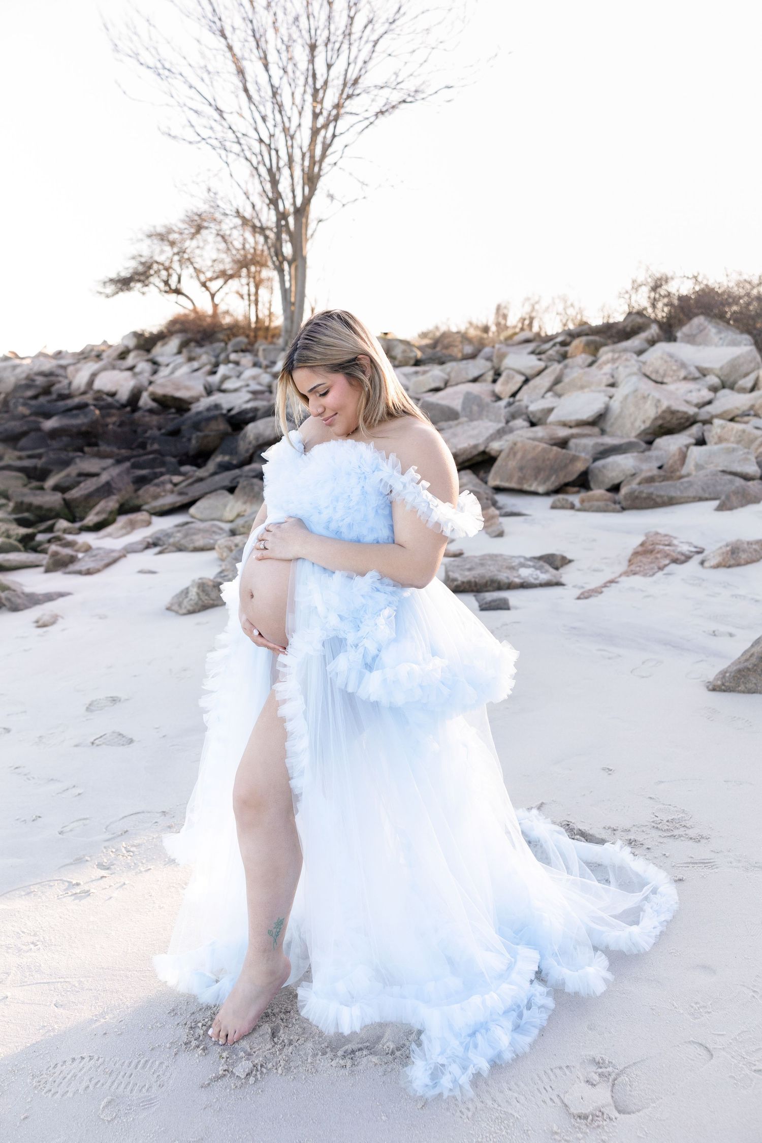 Beach maternity photos with ethereal light blue gown against rocky shore.