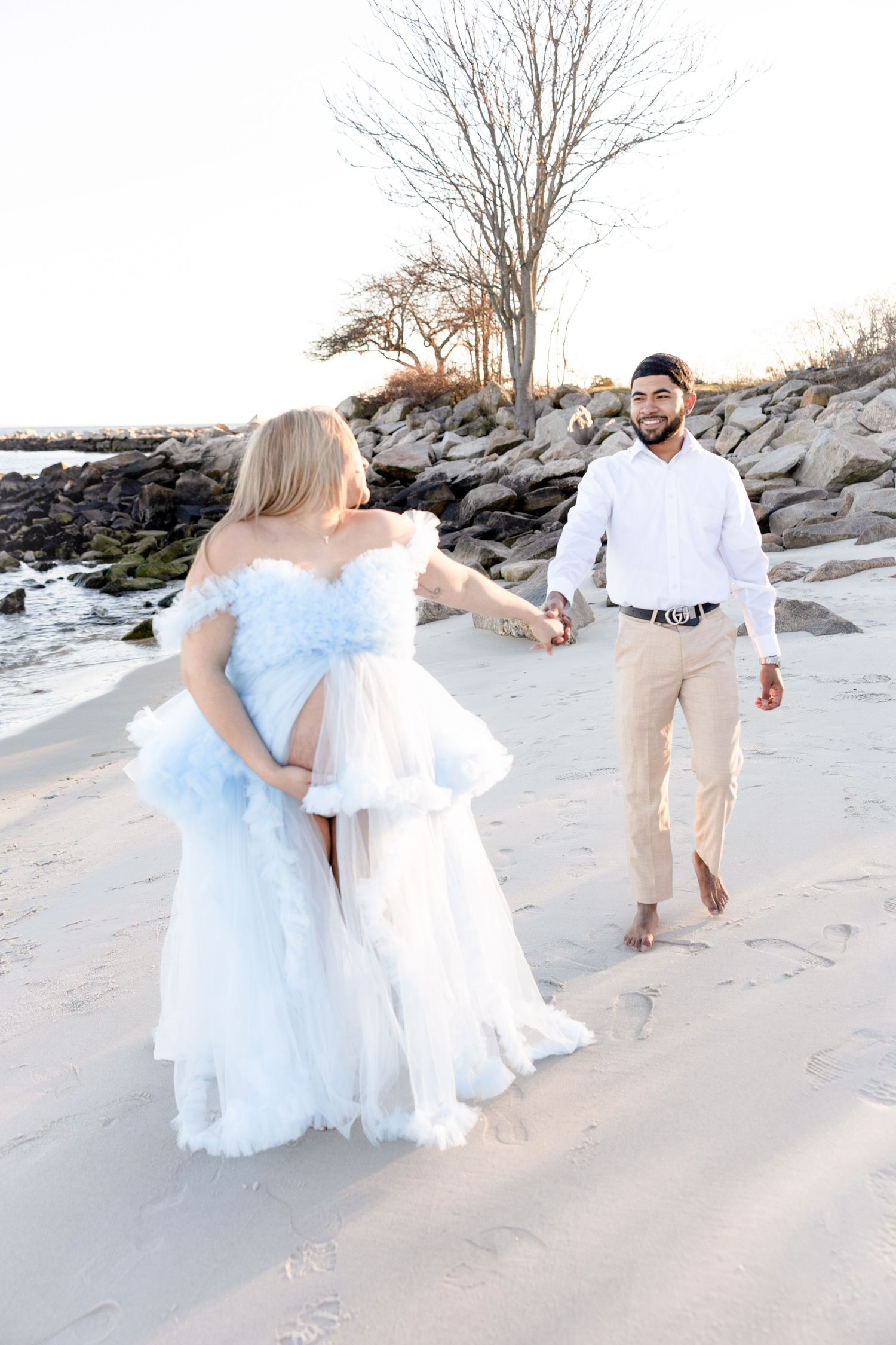 Couple walks along beach, woman in flowing light blue dress during sunset photoshoot.