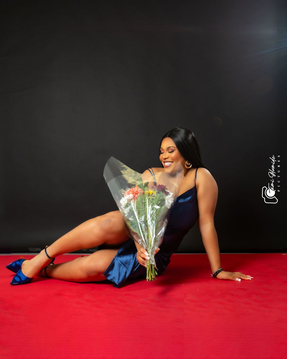 A glamour photoshoot featuring a navy blue dress and floral bouquet on a red backdrop.